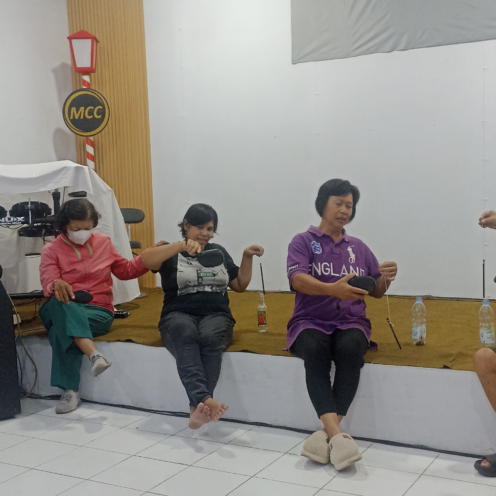 In Magelang, Indonesia, three women are seated on a raised platform in what appears to be an indoor event space. To the left, a woman wearing a pink jacket and green pants sits with a face mask on, holding a black object. In the center, a woman in a black t-shirt and dark pants is engaged in an activity involving a bottle, a string, and a stick. To the right, another woman in a purple polo shirt with "ENGLAND" written on it and black pants is also handling a string and a black object. Behind them, a decorative lamp post with an "MCC" sign is visible against a ribbed wooden wall. The floor is tiled white. There are several bottles of water on the platform.