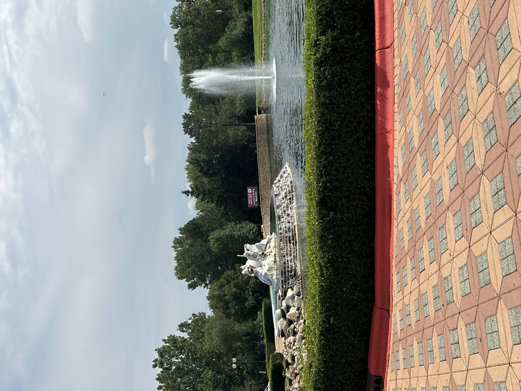 An outdoor scene features a large fountain with a sculpture of figures in a pond. The fountain sprays water high into the air. In the foreground, there is a decorative brick pathway with a red curb and a manicured hedge. Behind the pond, trees form a dense backdrop. A sign with red lettering and white text is visible on the far left, and a distant airplane is in the sky. The location is identified as the City of Bartlett, United States of America.