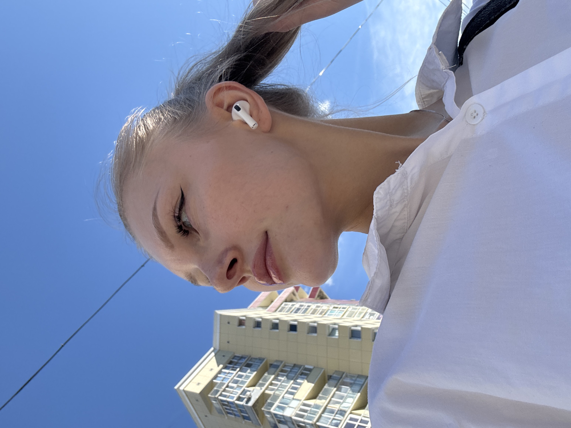 The media file is a close-up, low-angle selfie of a young woman outdoors, looking downwards. She has blonde hair pulled back and is wearing a white shirt with a collar and a black accent. A white wireless earbud is visible in her right ear. Her makeup includes eyeliner and lip gloss.
The background is dominated by a bright blue sky, suggesting it is daytime and the weather is clear. In the lower portion of the image, a tall, multi-story building with a cream-colored facade and numerous windows is visible. The building's architecture suggests an urban environment. A power line runs diagonally across the sky.
The woman's expression is neutral, and the overall mood is casual and relaxed. The bright sunlight indicates it is likely midday or afternoon. The location context provided is Perm, Russia. No visible text is present in the image.