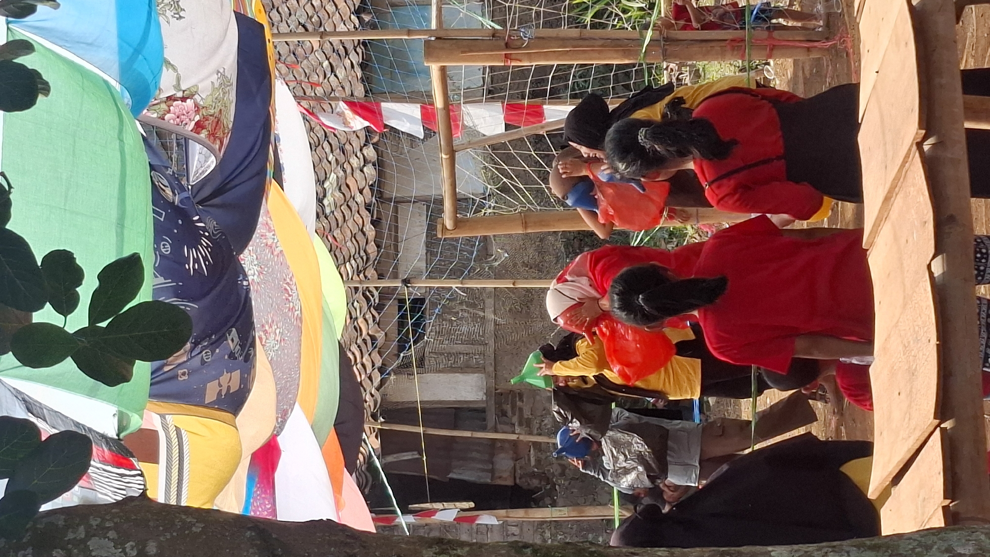 The image depicts an outdoor gathering in Cilaku, Indonesia, possibly a community event or celebration. The scene is partially covered by a colorful canopy made of various fabrics, creating a festive atmosphere. A group of people, including adults and children, are engaged in what appears to be a game or activity. Some individuals are holding large plastic funnels over their heads, while others are holding red buckets. The background shows traditional Indonesian houses with tiled roofs and woven walls. A net is set up, possibly for a game. The overall impression is one of lightheartedness and community participation, with elements of Indonesian culture visible in the surroundings and attire.