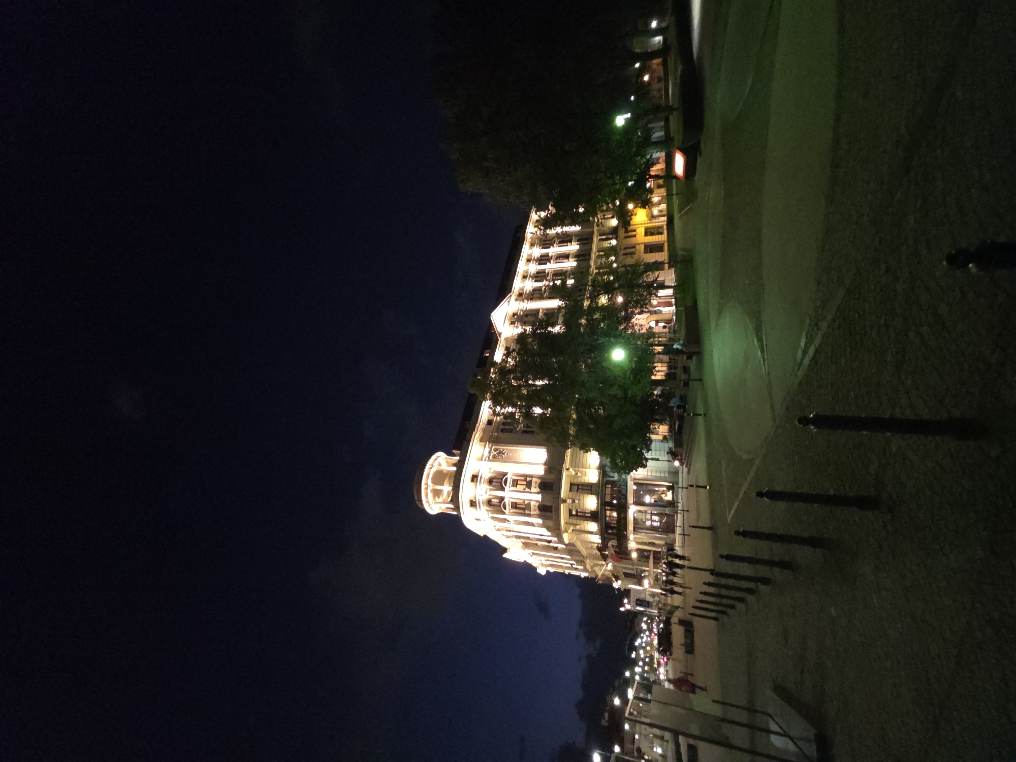 Captured at night in Warsaw, Poland, the image presents a brightly lit building, likely a hotel, prominently featured on the right side of the frame. This building stands in contrast to the dark sky that dominates the left side.
The foreground shows a paved walkway or plaza, marked by a series of dark, thin bollards. The illuminated facade of the building is lined with windows and decorative architectural details. Trees obscure some parts of the building. People are visible near the base of the building. Streetlights illuminate the foreground. The environment is urban, with a stark contrast between the bright, artificial light and the natural darkness.