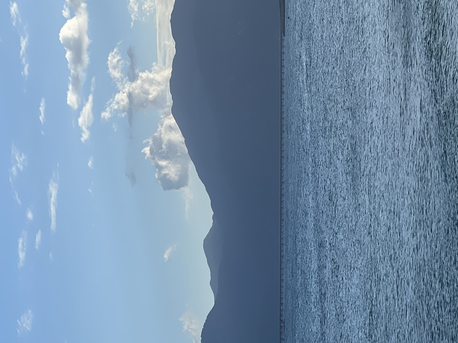 The image captures a coastal landscape in Ninh Hòa, Vietnam. A pale blue sky occupies the upper third of the frame, featuring scattered white cumulus clouds, with a larger cluster positioned above the central mountains. A dark, silhouetted mountain range forms the middle ground, extending horizontally across the scene. Below the mountains, a long, slender pier or bridge structure, supported by numerous vertical pilings, traverses the body of water. The lower two-thirds of the image consist of rippling blue-green water, presenting a textured surface. No human figures, animals, or active objects are present within the depicted environment.