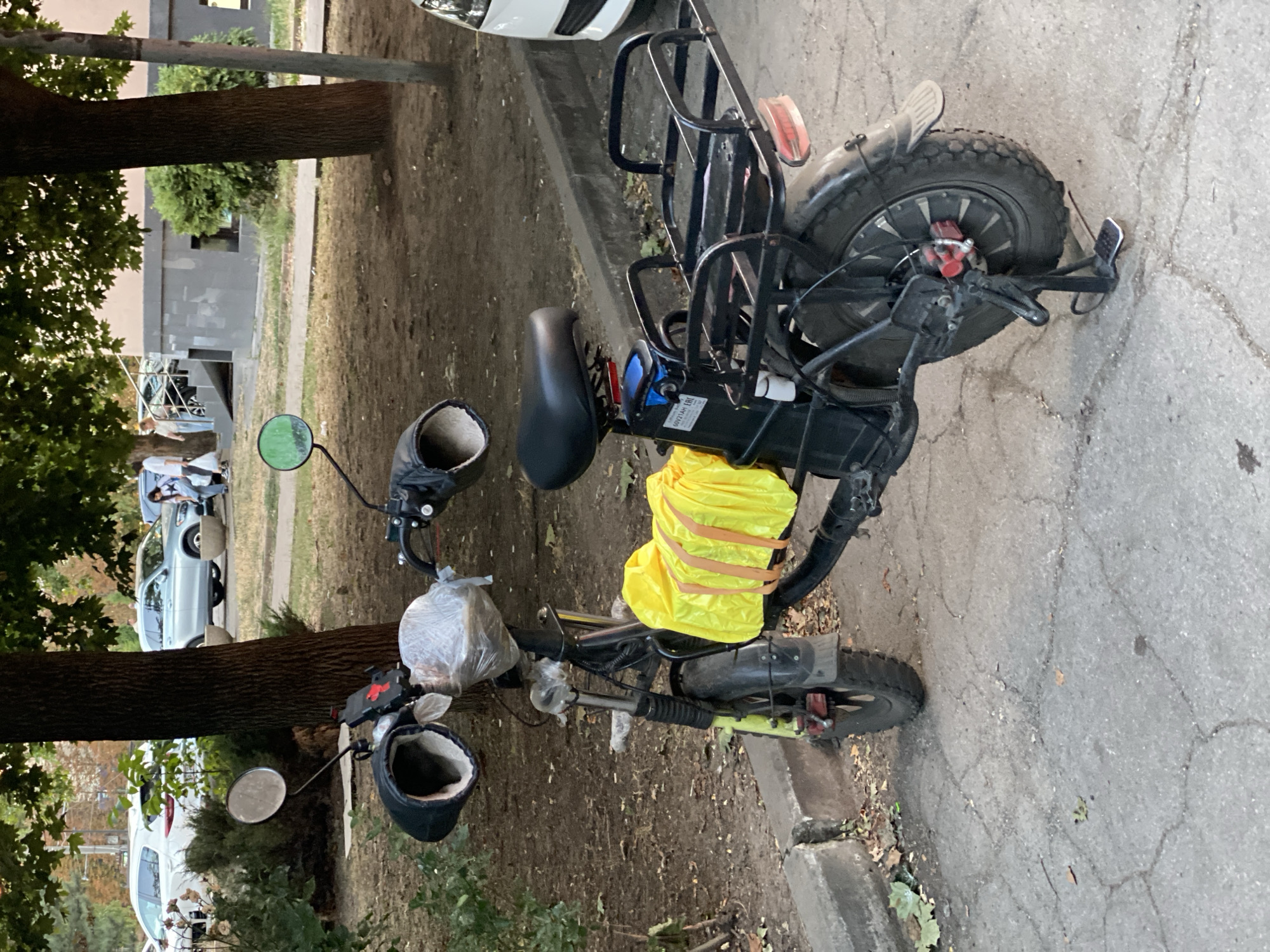 A black electric scooter is parked on a cracked asphalt surface next to a curb in a residential area. The scooter has a black seat, a rear rack, and a bright yellow bag secured with straps to the frame. The front of the scooter has two hand warmers and a clear plastic bag covering part of the handlebars. In the background, there are trees, a building with a grey facade, and several parked cars, including a silver SUV and a white car. Two individuals are visible in the background walking towards the building. The environment appears to be a city street or sidewalk in Chișinău, Moldova, based on the available context.
