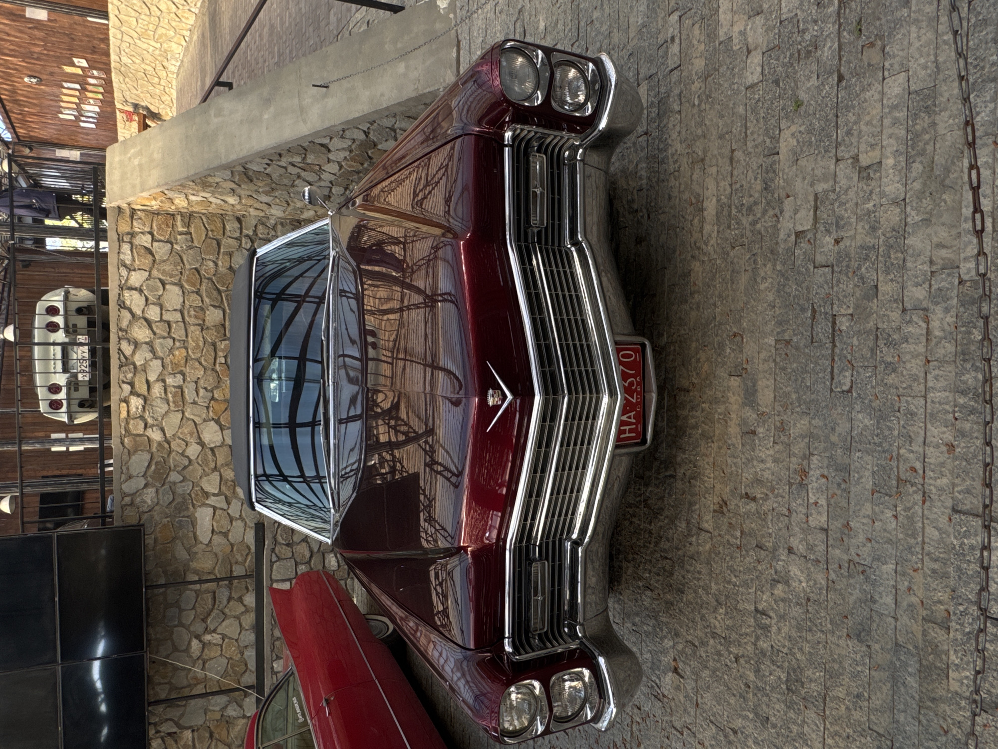 The image showcases a vintage burgundy Cadillac, potentially a 1967 model, parked on a stone-paved surface. The car's front grill is prominent, and a license plate that reads "HA-2370 Cuba" is visible. The car is surrounded by stone walls and metal fencing, suggesting an outdoor or semi-outdoor display or parking area. The location may be a car museum or vintage car dealership. There is no indication of the location in the city of город Ялта, Ukraine.