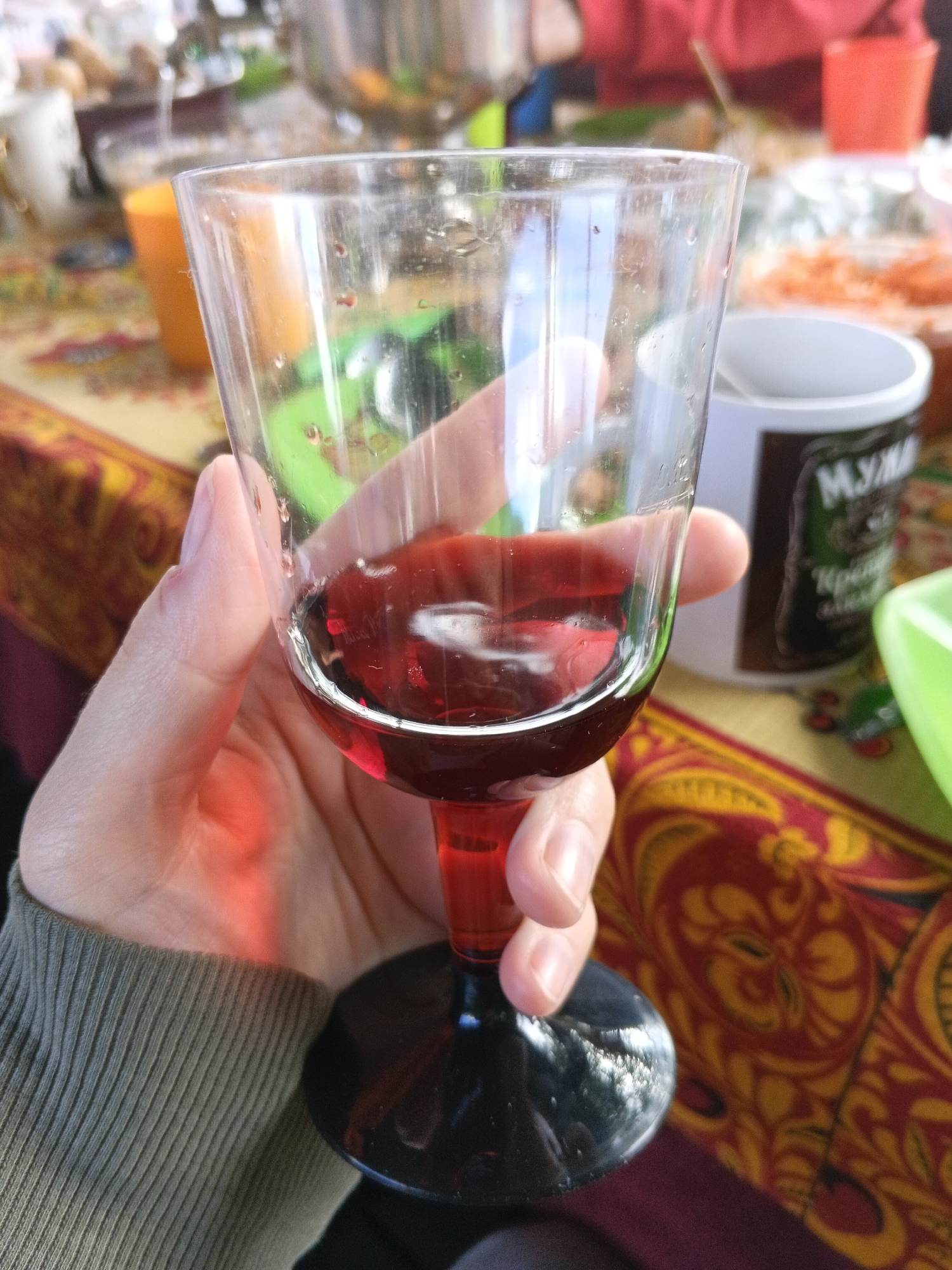 A person's hand, with visible skin texture and fingernails, is holding a clear glass goblet filled with a red liquid, presumably wine. The goblet has a red stem and a black base. The glass is slightly smudged with water droplets. In the background, a table is covered with a patterned tablecloth featuring red and yellow floral designs. Various objects are present, including an orange cup, a white mug with dark lettering that appears to be Russian, and a green container. The scene suggests an informal gathering, possibly outdoors or in a brightly lit room, given the reflections and lighting on the glass. There is no discernible indication of the city of Medvedevka, Russia, within the image itself.