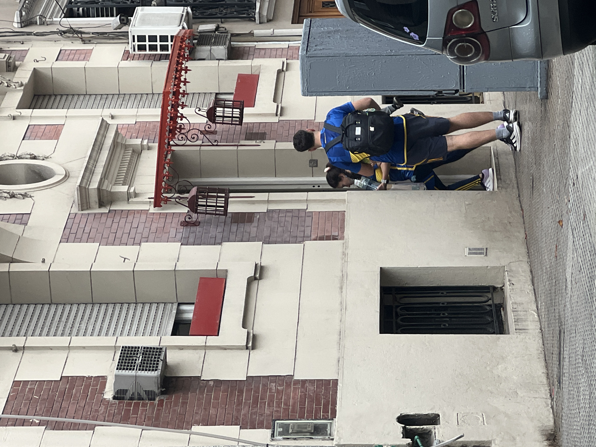 Two people are bent over in front of a building, with one wearing a blue shirt and a backpack. The building's facade is characterized by a mix of beige and brick sections, adorned with red awnings and decorative metalwork. A silver car is partially visible at the top of the frame, positioned near the building. The setting appears to be an urban environment. The image was likely taken in Buenos Aires, Argentina, given the architectural style of the building.