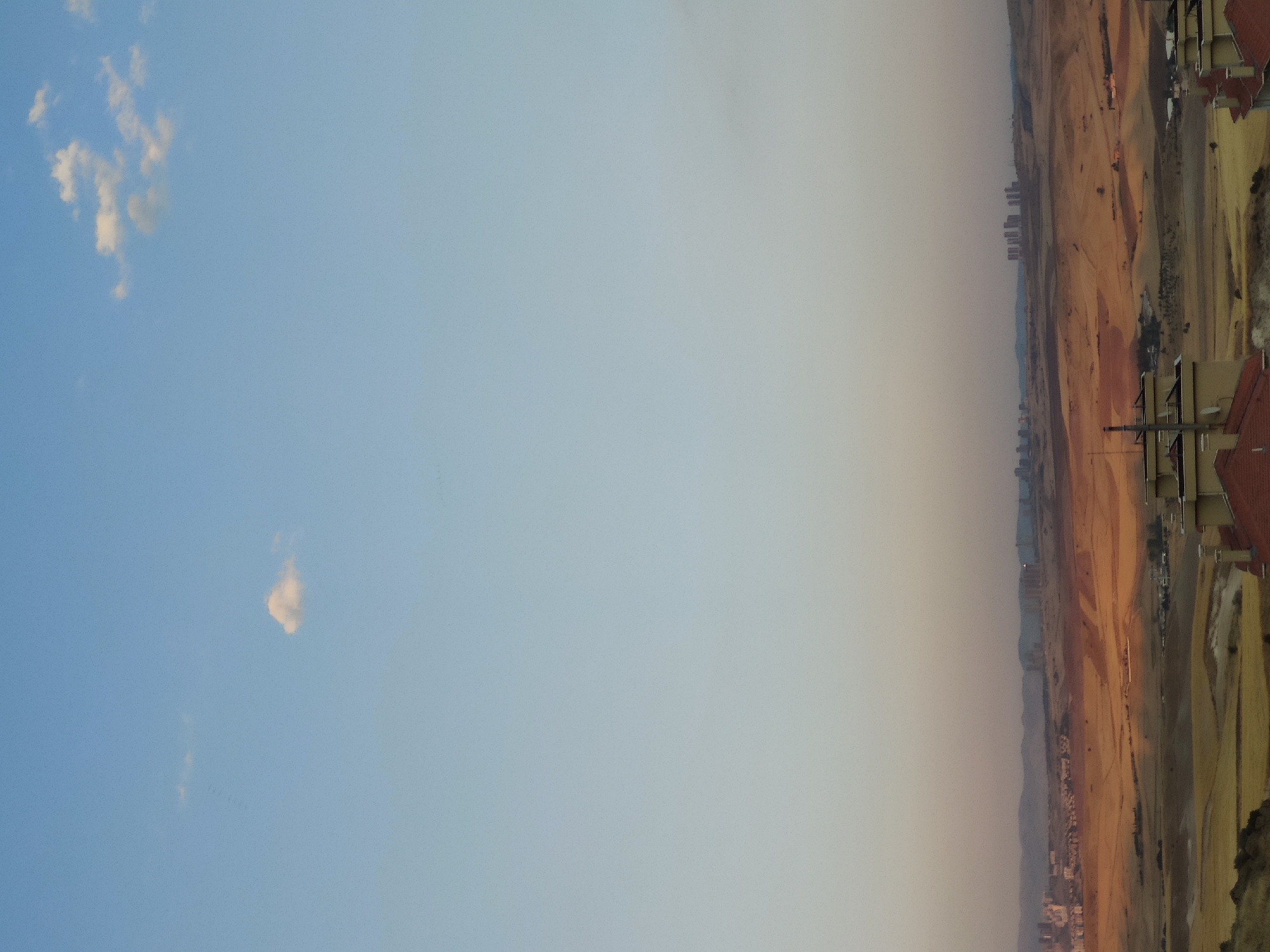 The image depicts a panoramic view of the outskirts of Etimesgut, Turkey, likely taken during the late afternoon or early evening. The sky is a soft gradient of pale blue to a muted orange-grey near the horizon, with a few scattered white clouds. In the foreground, the tops of residential buildings with red-tiled roofs are visible.
The middle ground reveals a vast, undulating landscape characterized by arid, yellowish-brown terrain interspersed with patches of darker, possibly cultivated fields. The terrain suggests a semi-arid climate. Distant cityscapes are visible on the horizon, featuring clusters of modern buildings and skyscrapers, indicating the presence of urban development in the area. The overall scene conveys a sense of open space and the transition between rural and urban environments.