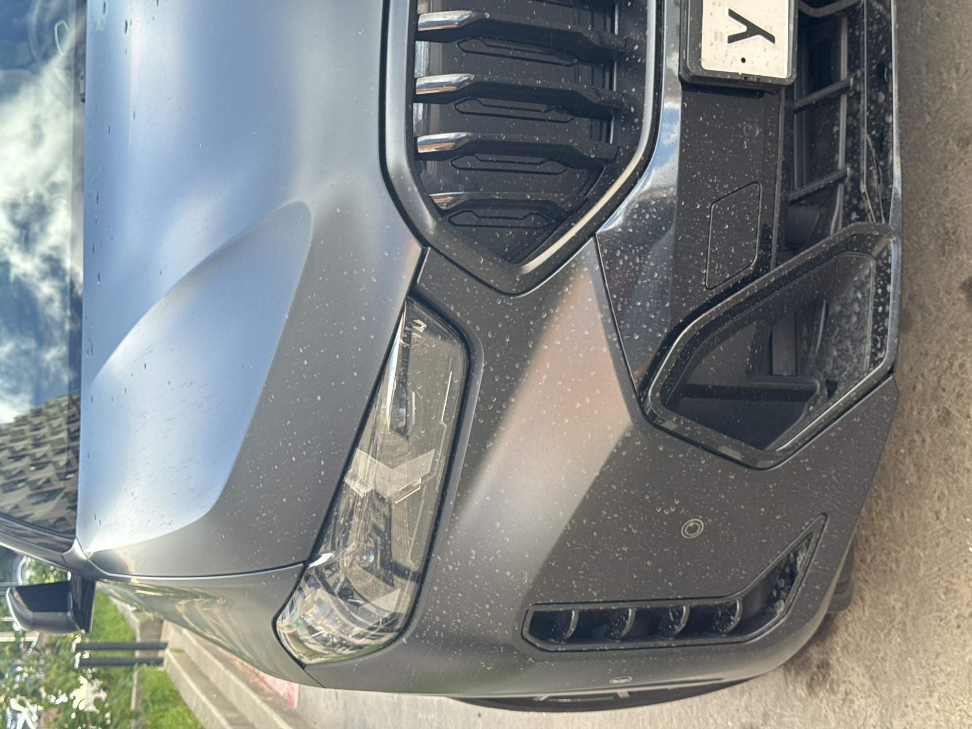 The image shows the front passenger side of a dark gray, matte-finished BMW X6. The vehicle is covered in a thin layer of dust and water spots. The license plate is visible, with the Cyrillic letter "У" and a Russian Federation flag. The car is parked on a paved surface, likely a street or parking lot, with a concrete curb and green foliage to the left. In the background, a modern building with reflective windows is visible. The scene appears to be in an urban environment in Moscow, Russia.