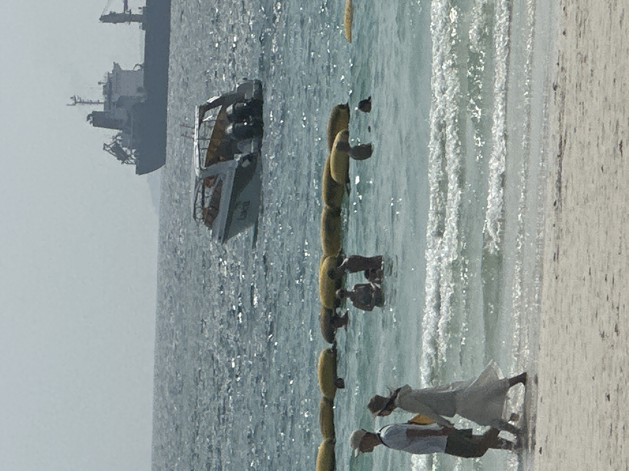 A horizontal eye-level shot depicts a beach scene, likely in Pattaya City, Thailand. A tan sand beach is on the right. A couple is seen walking on the beach toward the viewer. A person in a dress and hat is walking beside a person wearing a hat and a light-colored shirt and dark shorts. In the center of the image is an ocean with small waves, a yellow plastic barrier, and several people knee-deep in the water near the plastic barrier. Further out is a small motorboat and a very large ship. The sky appears overcast.