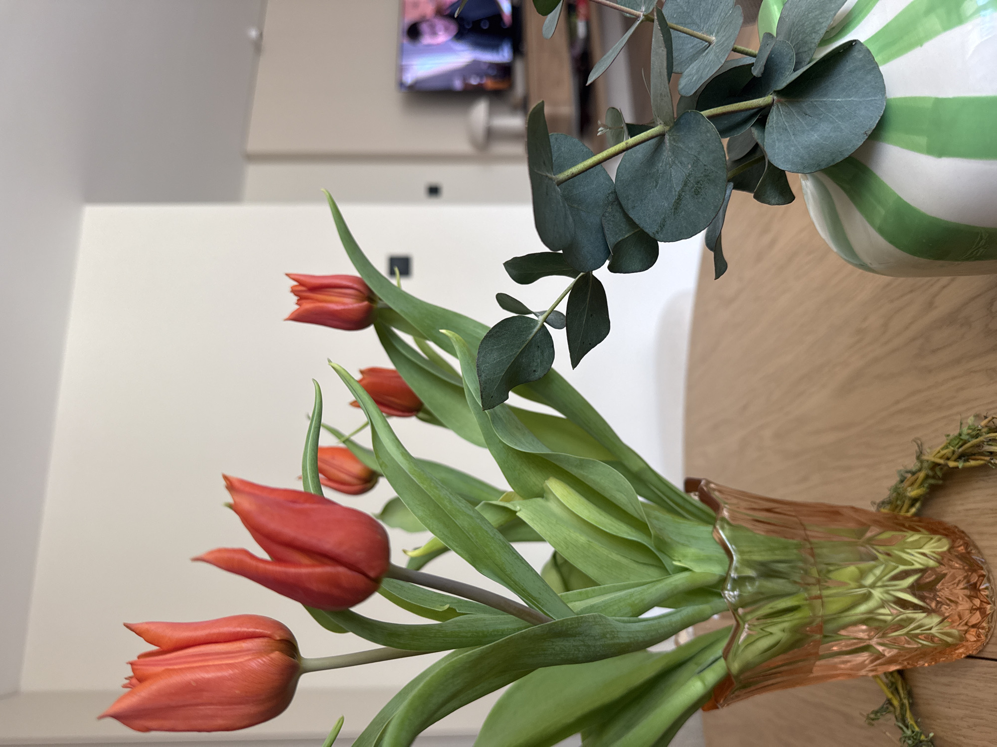 The image depicts a bouquet of red tulips and eucalyptus leaves in an orange, patterned vase. The vase sits on a wooden surface. In the background, out of focus, is a white wall with a television mounted on it. There are no people visible in the frame. It is not possible to ascertain whether the location is in the city of Burgas, Bulgaria.