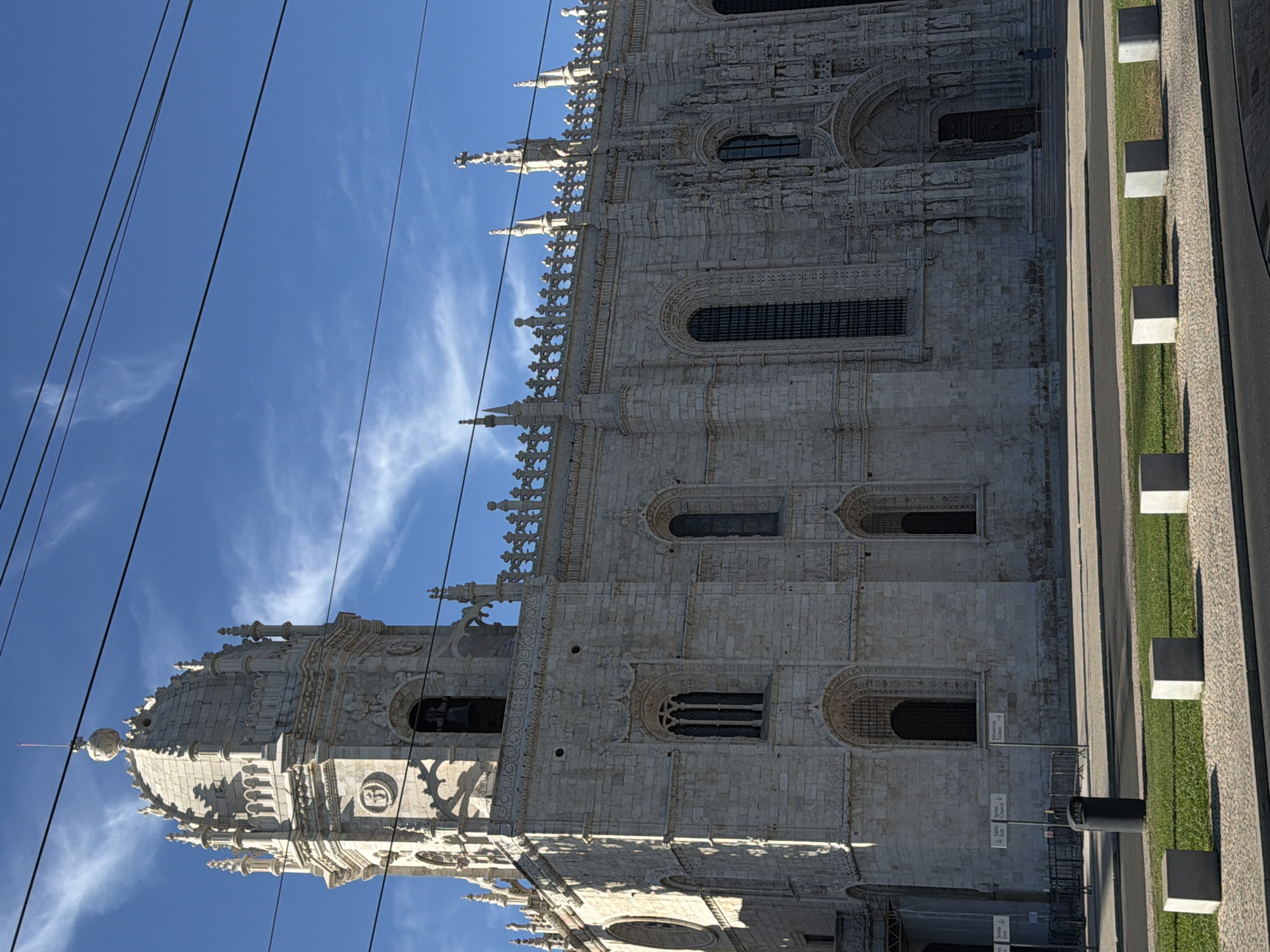 The Jerónimos Monastery, a UNESCO World Heritage site located in the Belém district of Lisbon, Portugal, is depicted in this image under a bright blue sky with wispy clouds. The monastery's elaborate Manueline architecture, characterized by its ornate stone carvings and pointed arches, is prominently displayed. The left side of the building features a tall, multi-tiered tower topped with a dome and finial. Numerous windows, some arched and others rectangular, punctuate the stone facade. Decorative crenellations and spires adorn the roofline. In the foreground, overhead wires are visible against the sky. A paved walkway runs in front of the monastery, with a strip of green grass and a line of concrete barriers separating it from a road. A trash bin and informational signs are also present. The scene is devoid of any visible action or interaction among the few distant figures that are present.