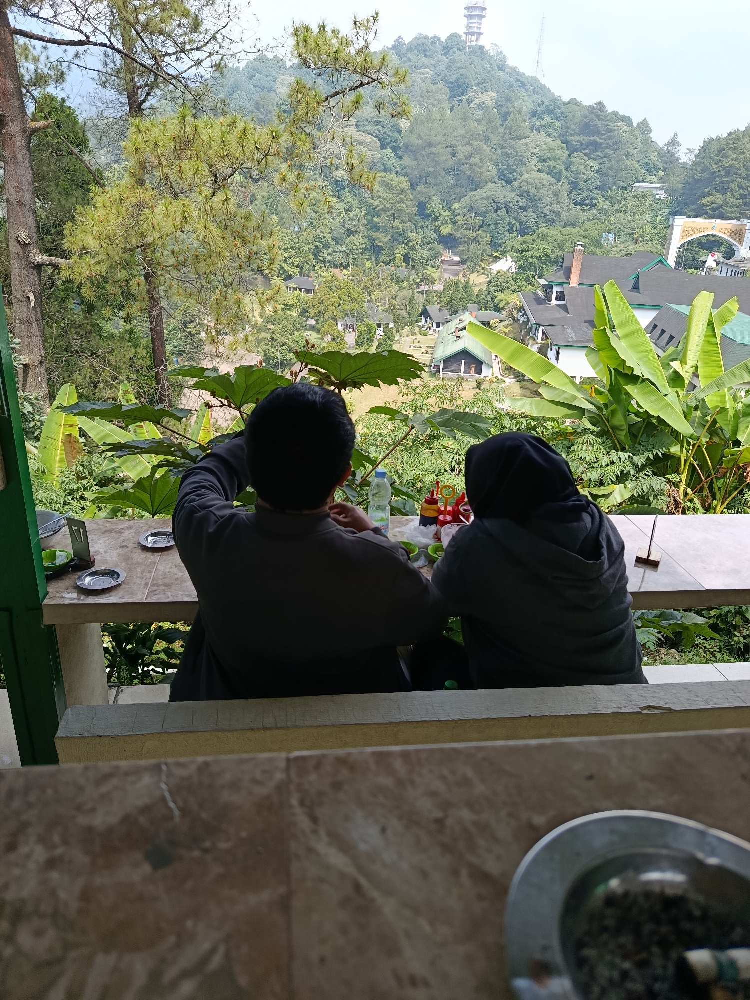 The image depicts two individuals from behind, seated at a table on a balcony overlooking a lush, green landscape. The individuals appear to be a man wearing a dark gray shirt and a woman wearing a dark hijab and gray jacket. They are facing away from the camera and towards the scenic view. On the table in front of them are several items, including what appears to be a bottle of water, three small condiment bottles (likely chili sauce, ketchup, and soy sauce), and two small ashtrays. One ashtray contains cigarette butts. The table itself is made of light-colored tiles and supported by a concrete pillar.
The background reveals a dense forest covering rolling hills. Several buildings with green roofs are visible nestled within the trees, suggesting a residential or resort area. In the distance, atop one of the hills, a tall tower structure can be seen. The foliage in the foreground includes large, broad leaves and pine trees.
The overall scene suggests a relaxed outdoor setting, possibly a cafe or a home balcony, offering a panoramic view of the natural surroundings. The presence of condiments and ashtrays indicates that the individuals may have been eating or smoking. The location is likely a mountainous or hilly region, given the extensive tree cover and the distant tower structure. The mention of Cianjur, Indonesia, places this scene within a region known for its natural beauty and cooler climate due to its elevation.