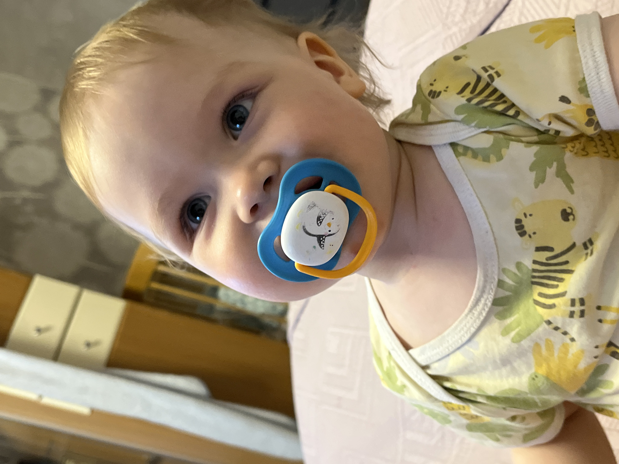 A close-up shot shows a baby with fair skin and blonde hair, lying on a surface that appears to be a bed, given the light purple color and texture. The baby has a blue and yellow pacifier in its mouth. The baby is wearing a light yellow and green patterned shirt. In the background is furniture, possibly drawers with silver knobs. The location is not discernible from the image, therefore, it is not possible to pinpoint it as being in Chaikovsky, Russia.
