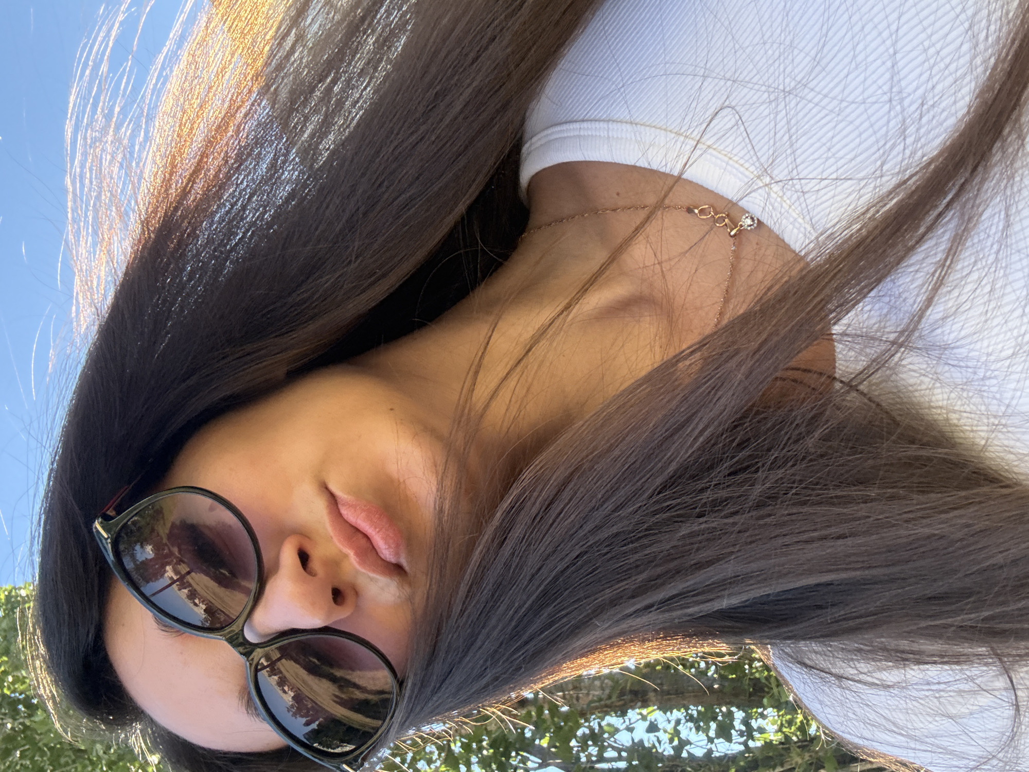A close-up, low-angle selfie shows a woman from the chest up, outdoors. The woman has long, dark brown hair that is partially blowing across her face. She is wearing dark-rimmed sunglasses, a white ribbed shirt, and a thin gold chain with a small pendant. Her lips are slightly parted and painted with a neutral shade of lipstick. The background is a bright blue sky and out-of-focus green foliage. The image appears to have been taken in Kyiv, Ukraine.