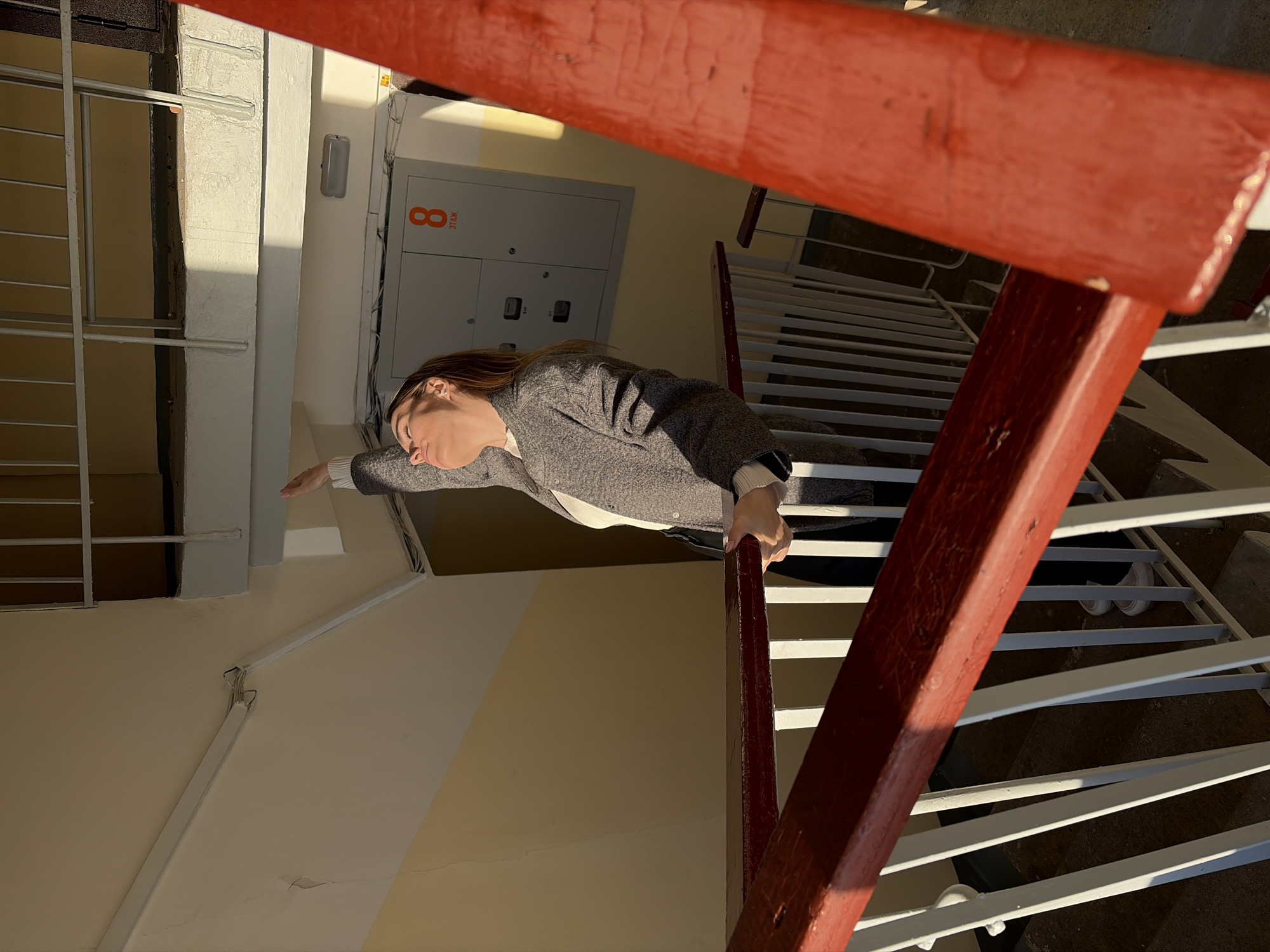 The image depicts a woman standing on a staircase in a building in Izhevsk, Russia. She is wearing a gray sweater and white shirt, with black pants. She is holding onto a red wooden railing. There is a light tan wall in the background and a metal panel labeled "8 этаж". The staircase has white metal railings and concrete stairs. The lighting suggests it is daytime.