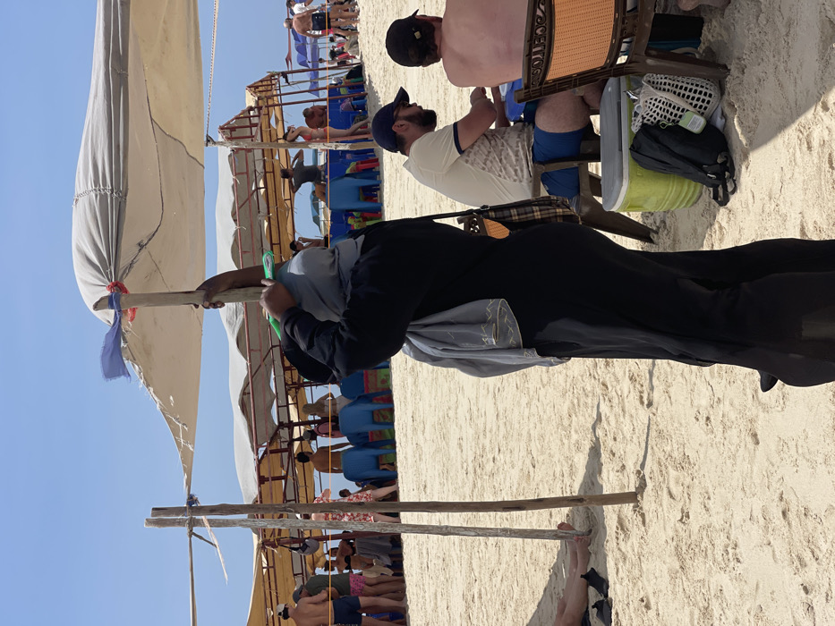The image depicts a bustling beach environment in Zanzibar, Tanzania, under a bright blue sky.
In the foreground, a person wearing a black full-length garment and a grey head covering stands with their back partially to the viewer, holding an upright wooden pole that supports a light-colored fabric canopy. A green clip is visible near their hands on the pole. A grey shawl with a gold patterned edge is draped over their left shoulder. To the immediate right, two men are seated on low stools. The closer man wears a light-colored patterned t-shirt, blue shorts, and a dark baseball cap. The further man is shirtless and wears a dark baseball cap. A green cooler, a black backpack, and a white mesh bag are on the sand near these seated men. To the left of the central figure, another wooden pole is planted in the sand, and two bare feet with black footwear are partially visible on the sand.
The midground features multiple makeshift shade structures, constructed from wooden poles and light-colored fabric canopies, under which various individuals are present. People are seated on blue and colorful plastic chairs or standing. On the left side of the midground, a group of individuals, predominantly men in swim trunks, are congregated. On the right side of the midground, a person in a red bikini top and dark bottoms is visible near another individual. The ground consists of light-colored sand throughout the visible area.