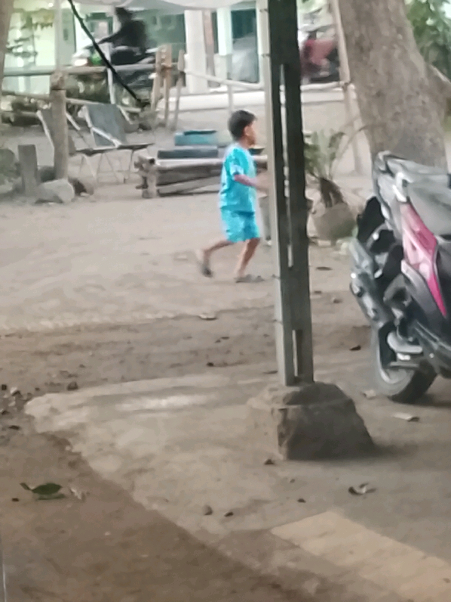 In Ujungrusi, Indonesia, a low-resolution, slightly blurred image depicts an outdoor scene with a young person in a bright blue t-shirt and shorts running across a dirt and gravel ground. The person appears to be moving from left to right within the frame. To the right of the running individual, a motorcycle with dark bodywork and a pink accent is parked. Behind the person and motorcycle, a large tree trunk is visible, along with some foliage and what appears to be a light-colored shelter or awning structure. In the background, there are wooden posts, some furniture including chairs, and debris scattered on the ground. The lighting suggests it is daytime.