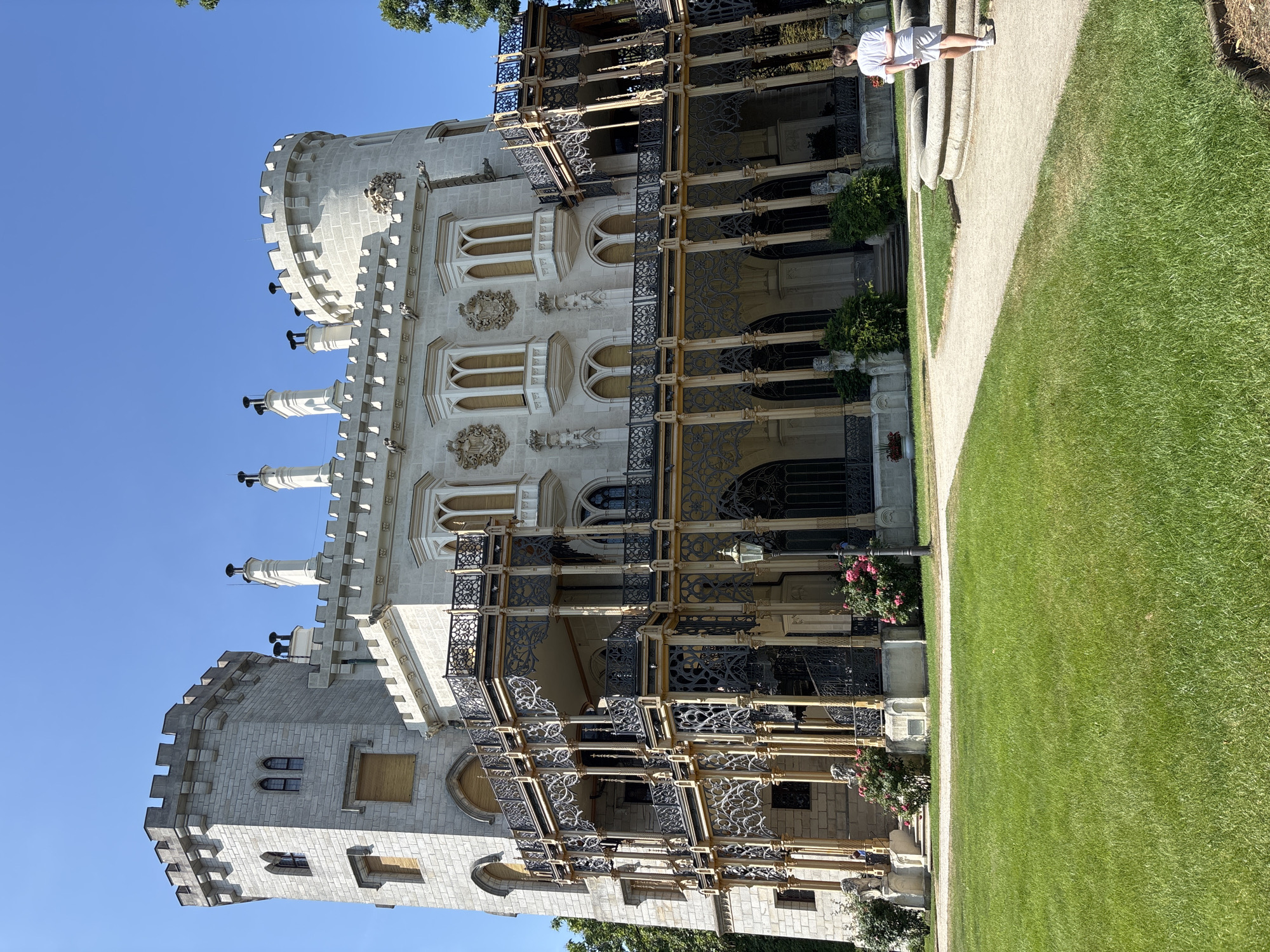 The image depicts Hluboká Castle, a Neo-Gothic landmark situated in Hluboká nad Vltavou, Czechia. The castle is characterized by its white stone facade, intricate ironwork balconies on multiple levels, and prominent turrets. Several tall, slender chimneys rise from the roofline. The castle is surrounded by a meticulously maintained green lawn, with a gravel path leading towards the building. A large, circular stone fountain is visible to the right of the path. A lone figure, a young person wearing a white t-shirt and shorts, walks away from the fountain on the path. Lush green trees are present on the right side of the frame, with some pink flowering bushes and a lamppost near the castle's entrance. The sky is a clear, bright blue.