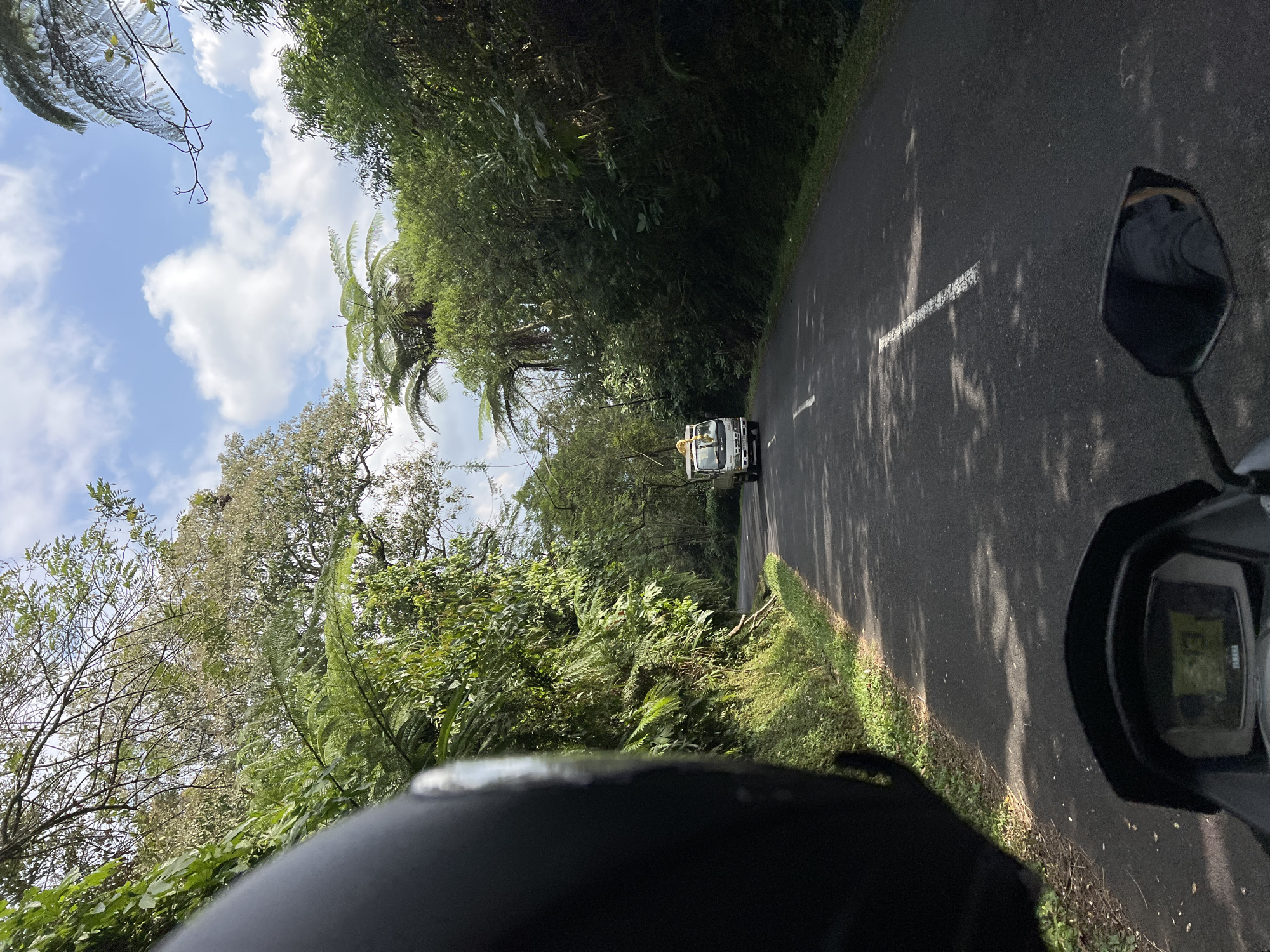A first-person perspective shot from a motorcycle looking forward along a paved road surrounded by lush green vegetation and trees. The road is empty except for a white truck in the distance, driving in the same direction. The sky is partly cloudy with a bright blue sky. The motorcycle's dashboard is visible in the foreground, displaying a speed of 13 km/h. The scene appears to be a rural area with dense foliage and winding road, consistent with locations in Pupuan, Indonesia.