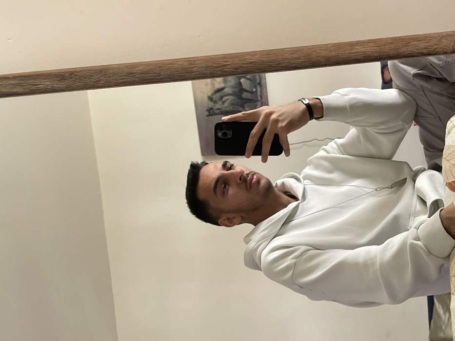 The scene takes place in an indoor residential setting, specified as being within the Neapoli Municipal Unit, Greece. A young adult male is captured in a mirror selfie, reflecting his upper body and head. He has short, dark hair and subtle facial hair, looking directly towards the smartphone in his right hand. He is wearing a light-colored, possibly off-white, hooded sweatshirt with a long-sleeved design, and a silver-toned necklace with an elongated pendant hangs around his neck. A dark wristband or watch is visible on his left wrist. The black smartphone, with a visible multi-lens camera module, is held vertically. The background, as seen in the mirror, features a plain light-colored wall. A framed artwork depicting two horses in muted tones is mounted on the wall to the subject's right. The mirror itself has a light brown, wood-grained frame, partially visible on the right side of the image. A section of a patterned fabric, possibly part of a bed or couch, is visible in the lower-left corner, suggesting the individual is seated.