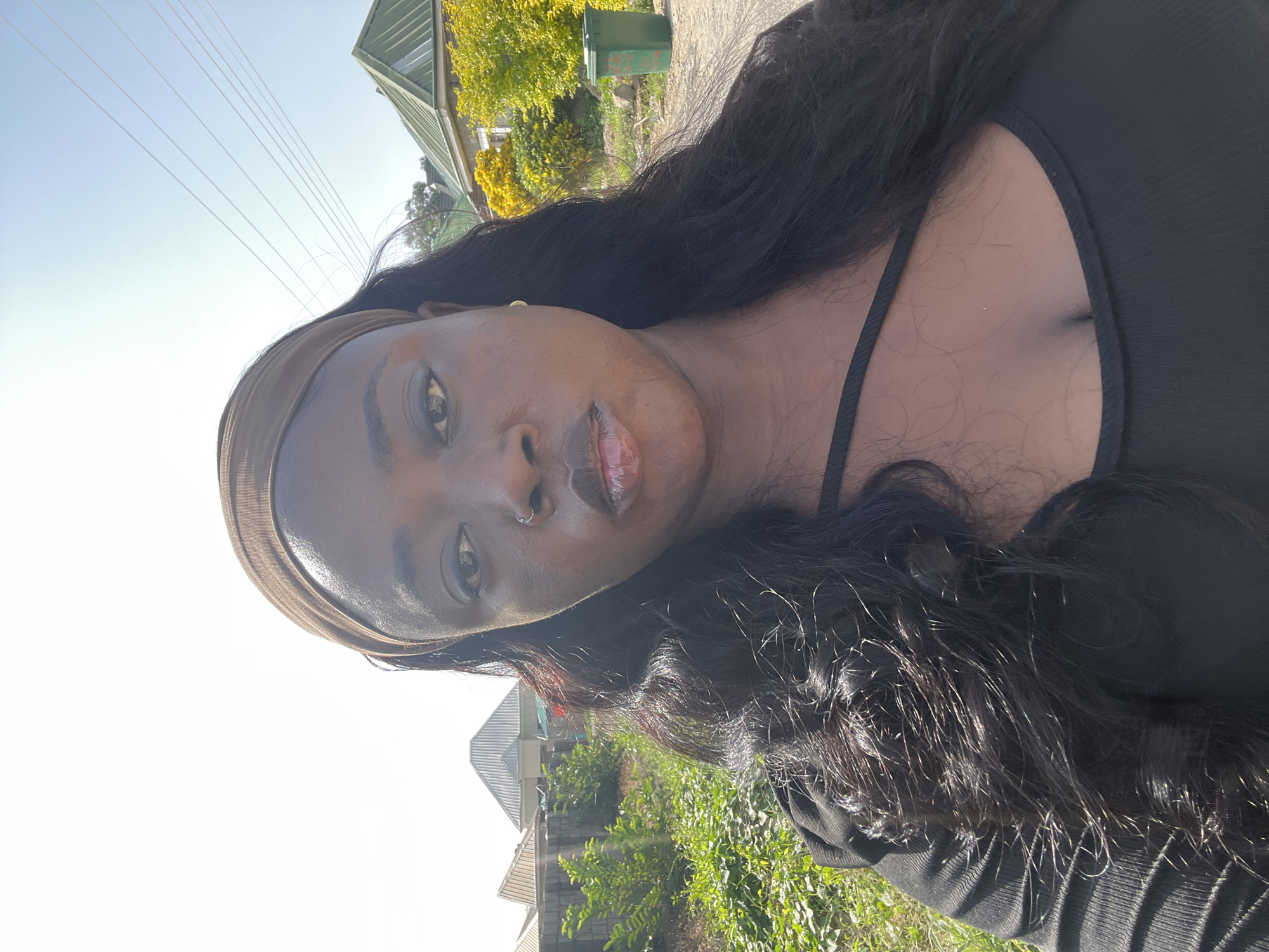 A woman with dark skin and long, dark hair is outdoors. She is wearing a light brown headband and a black top with thin straps. She has a nose ring and appears to be wearing lip gloss. The background includes houses with peaked roofs and greenery, likely in a residential area. The location is unspecified in the city of Abuja, Nigeria.