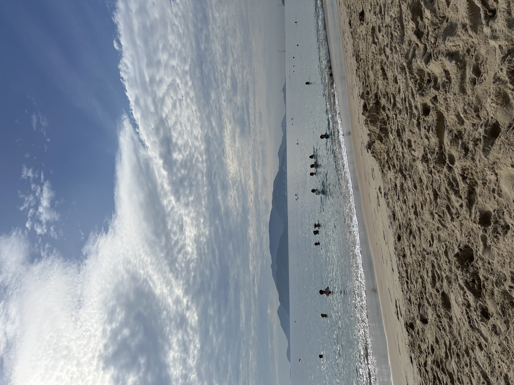 The image captures a beach scene in Nha Trang, Vietnam. The foreground is dominated by sand with visible footprints and small mounds, suggesting it's a well-used beach. The ocean occupies the middle ground, with a gentle wave line lapping at the shore. Several people are visible in the water, appearing to be swimming or wading. The middle distance features a body of water stretching towards a mountainous coastline. The sky above is a mix of blue and white clouds, with some areas appearing overcast. In the far distance, faint outlines of structures and possibly a bridge or transmission tower can be discerned on the horizon. The overall impression is a moderately populated beach on a day with dynamic cloud cover.