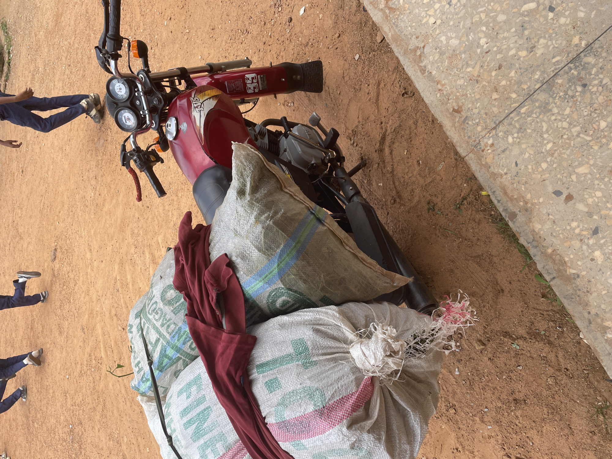 The image shows a red motorcycle parked on a dirt surface, with two large sacks tied to the back of it. The sacks are made of woven material, one is a light grey with green and blue stripes, and the other is white with green lettering and red and pink stripes. A dark red garment is draped over the sacks. In the background, parts of what appear to be people's legs and feet, dressed in blue trousers and casual shoes, are visible on the left side of the motorcycle. The scene is outdoors, with the ground being a mix of reddish-brown dirt and a concrete surface on the right side. The concrete appears to be a pathway or sidewalk, with small stones embedded in it. The lighting suggests it is daytime. The overall impression is that of a motorcycle being used to transport goods, possibly in a rural or semi-rural setting in Nigeria, given the context. There is no discernible text on the motorcycle itself, but the white sack has some visible green lettering.