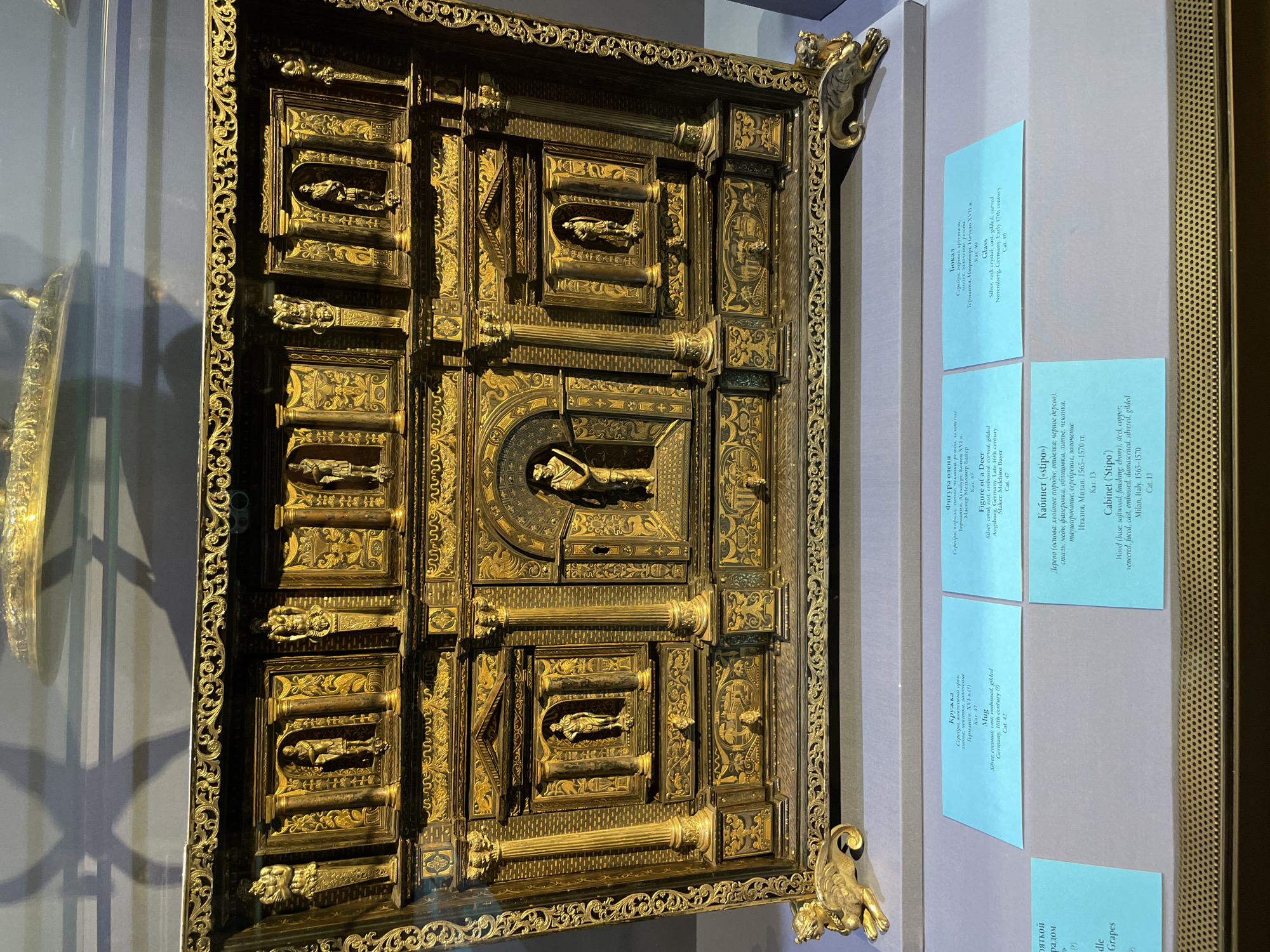 A close-up, slightly angled shot shows a square, highly ornamented gold-colored cabinet door, likely from a museum exhibit. The door is divided into multiple rectangular sections, each featuring relief sculptures of figures and decorative patterns. Small columns support the individual sections. The door is surrounded by an intricately detailed, ornate border with corner embellishments. To the right of the door, a display board with several blue informational cards is visible. The overall setting suggests an indoor museum environment, likely in an Unknown city.