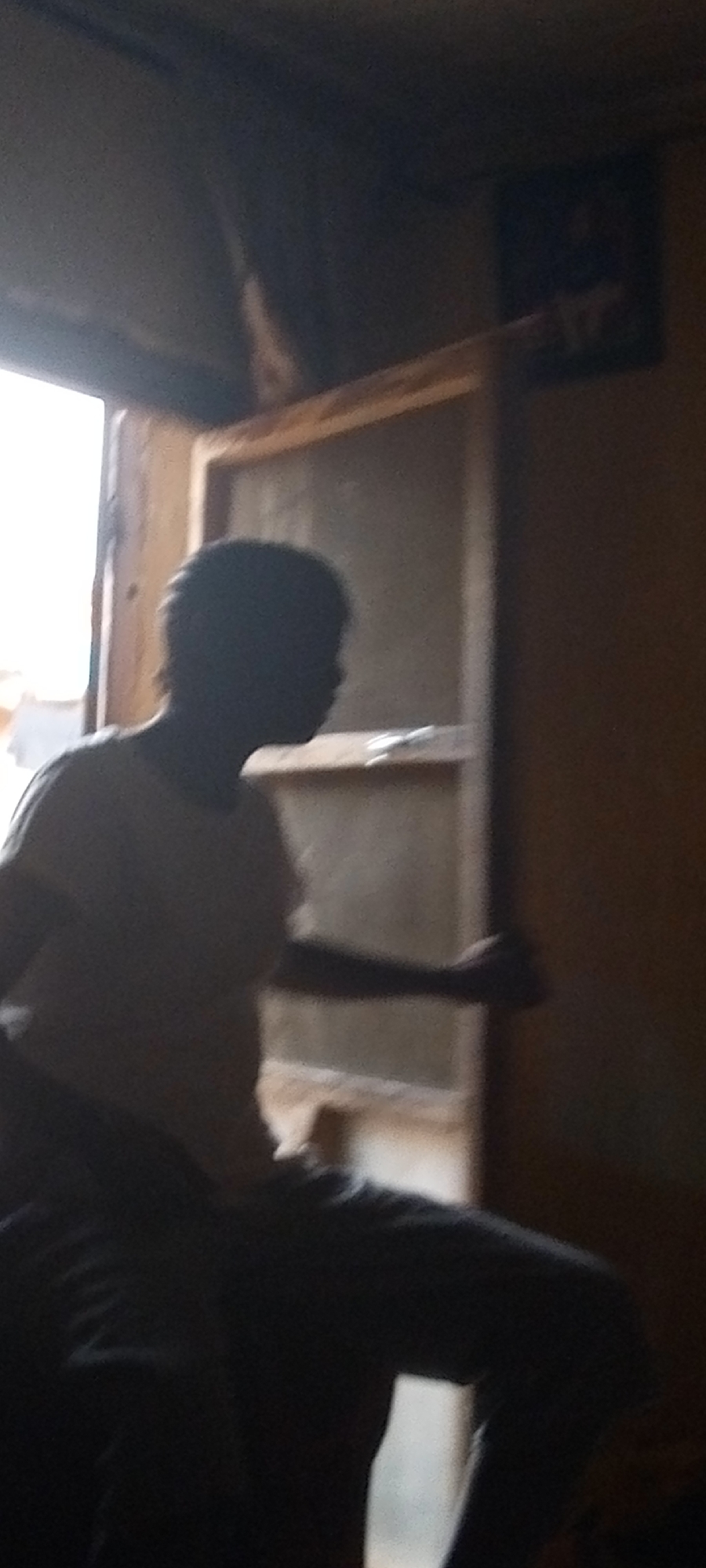 A person is standing in front of an open door. The location is a building in an unknown city. The person appears to be male, wearing a light-colored t-shirt and dark pants. The doorway leads to the outside. The environment seems dimly lit.
