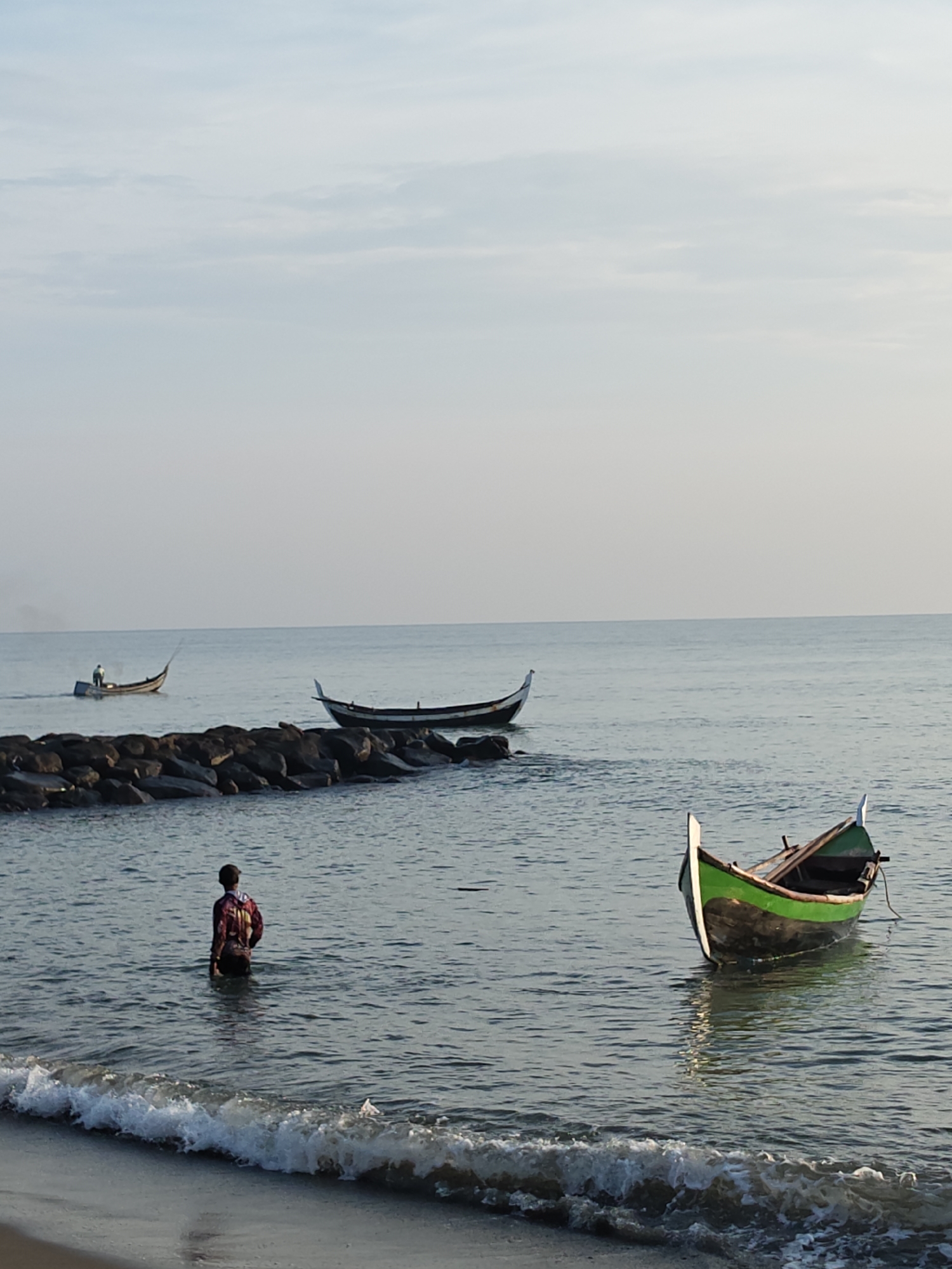 The image depicts a calm sea with a clear sky, suggesting it was taken during the day. In the foreground, waves gently lap against a sandy beach. A person is standing in the shallow water, facing away from the camera. The person appears to be wearing a colorful shirt.
Further out at sea, there are several boats. One boat, partially visible in the lower right corner, is green and brown. Another boat is positioned near a rocky breakwater, with a person inside. Two more boats are visible in the distance. The rocky breakwater extends into the water.
This scene is consistent with the coastal environment of Lhokseumawe, Indonesia, a city known for its beaches and fishing communities. The presence of traditional fishing boats and the calm sea are characteristic of the region.