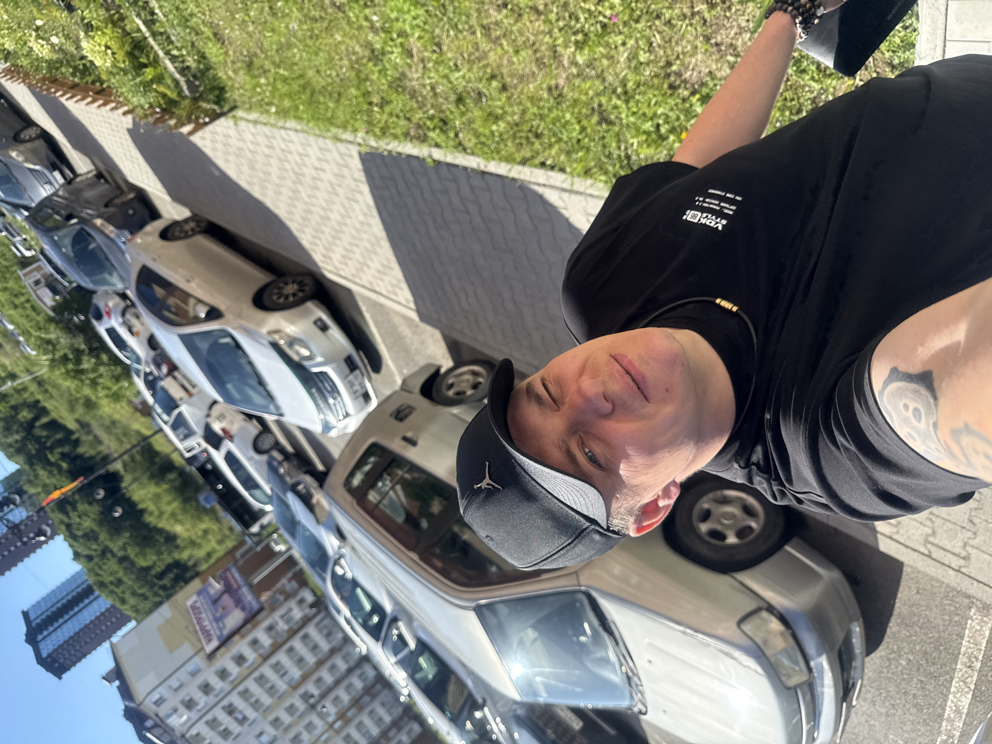 A man wearing a black baseball cap and a black t-shirt is standing in front of parked cars on a sunny day in Vladivostok, Russia. The man is in the foreground, with his face and upper body visible. He has a tattoo on his arm. Behind him, several cars are parked in a row. The cars are of various makes and models, and appear to be in a residential area. In the background, there are buildings and trees, suggesting an urban environment. A small patch of grass is visible on the right side of the image.