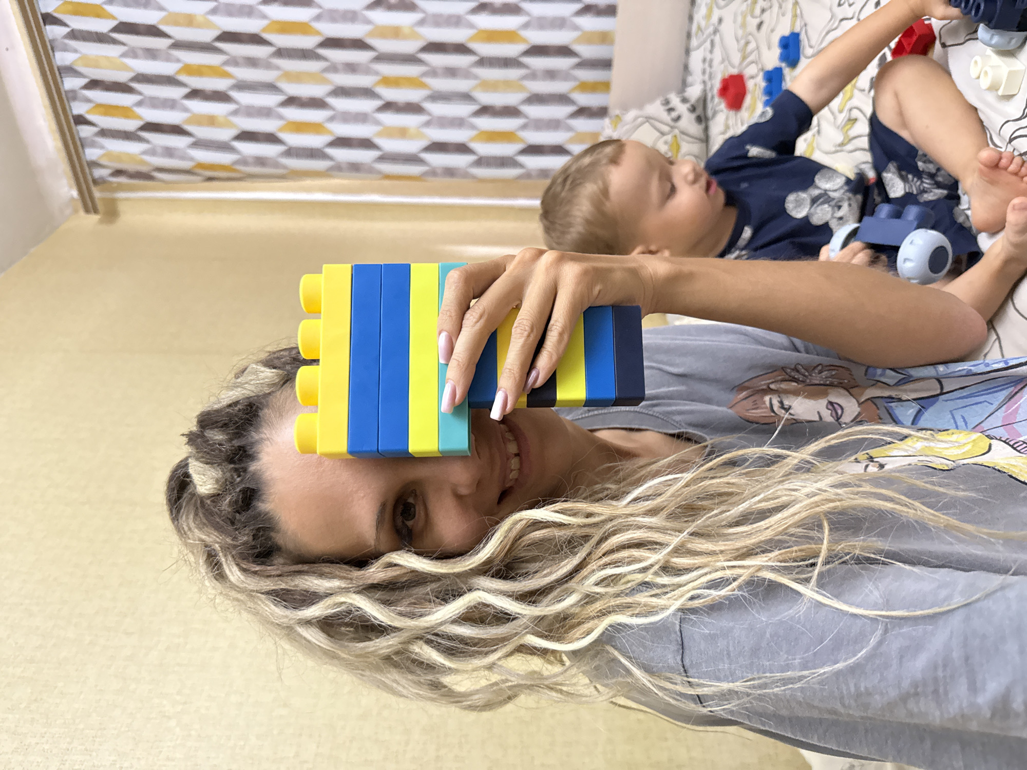A woman with blonde, curly hair braided into sections is holding a stack of blue and yellow building blocks in front of her face, obscuring her eyes. She is smiling and looking at the camera. She is wearing a grey t-shirt with a cartoon graphic. To the right and behind her, a young boy with brown hair is sitting on a bed. He is wearing a dark blue shirt with a Mickey Mouse print and is playing with building blocks. The bed is covered with a patterned duvet. In the background, a yellow wall and a curtain with a geometric pattern are visible. The scene appears to be an indoor setting, likely a bedroom. No specific location in Rusanovo, Russia, is depicted or inferable from the image.