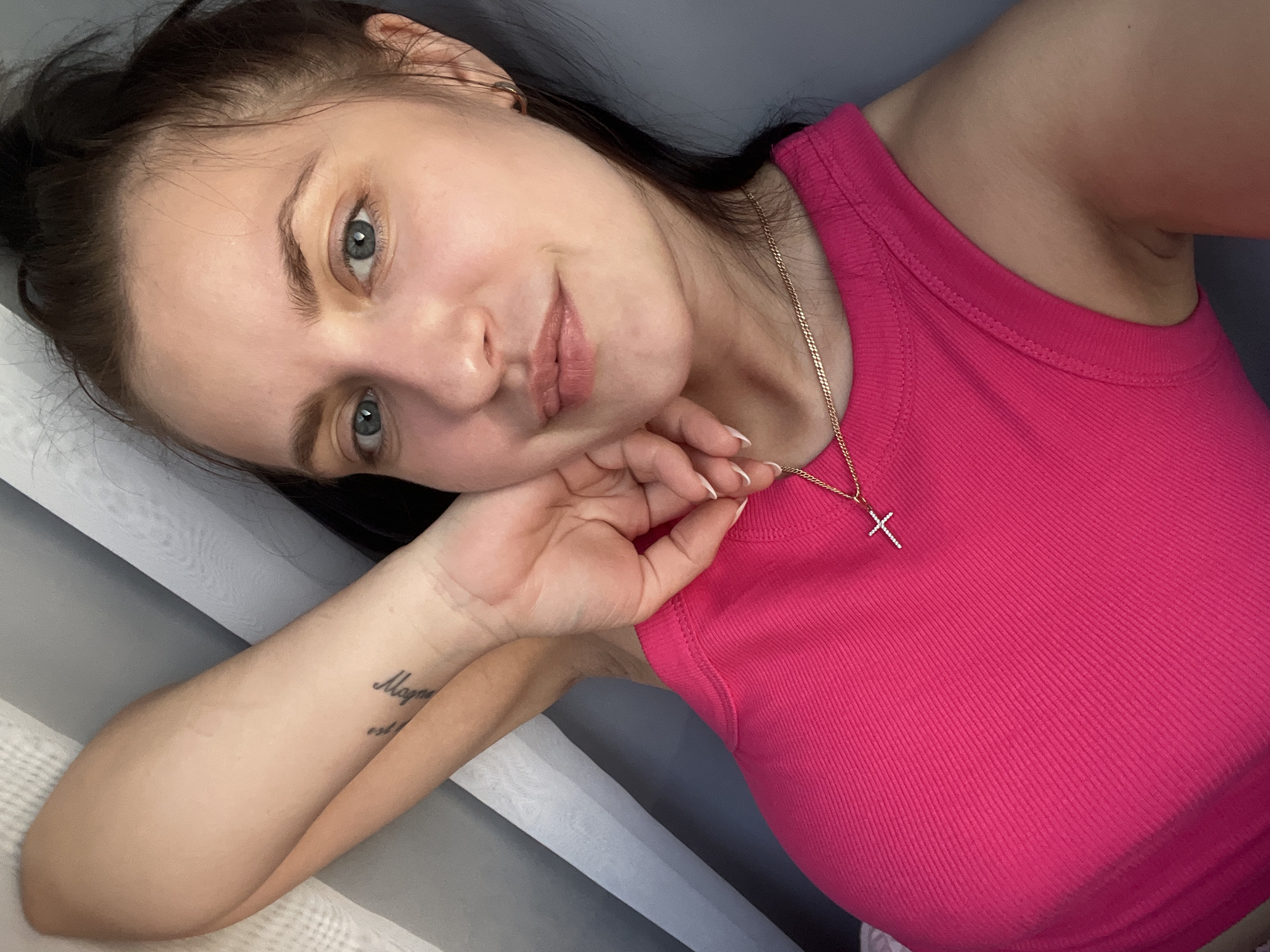 A woman with fair skin, light blue eyes, and dark hair poses in a selfie. She is wearing a bright pink tank top and a gold necklace with a cross pendant. Her left hand is raised to her face, with her fingers gently touching her cheek. A tattoo is visible on her inner forearm. The background features a window with sheer white curtains and a gray wall. The location is possibly in the city of Марковский, Russia.