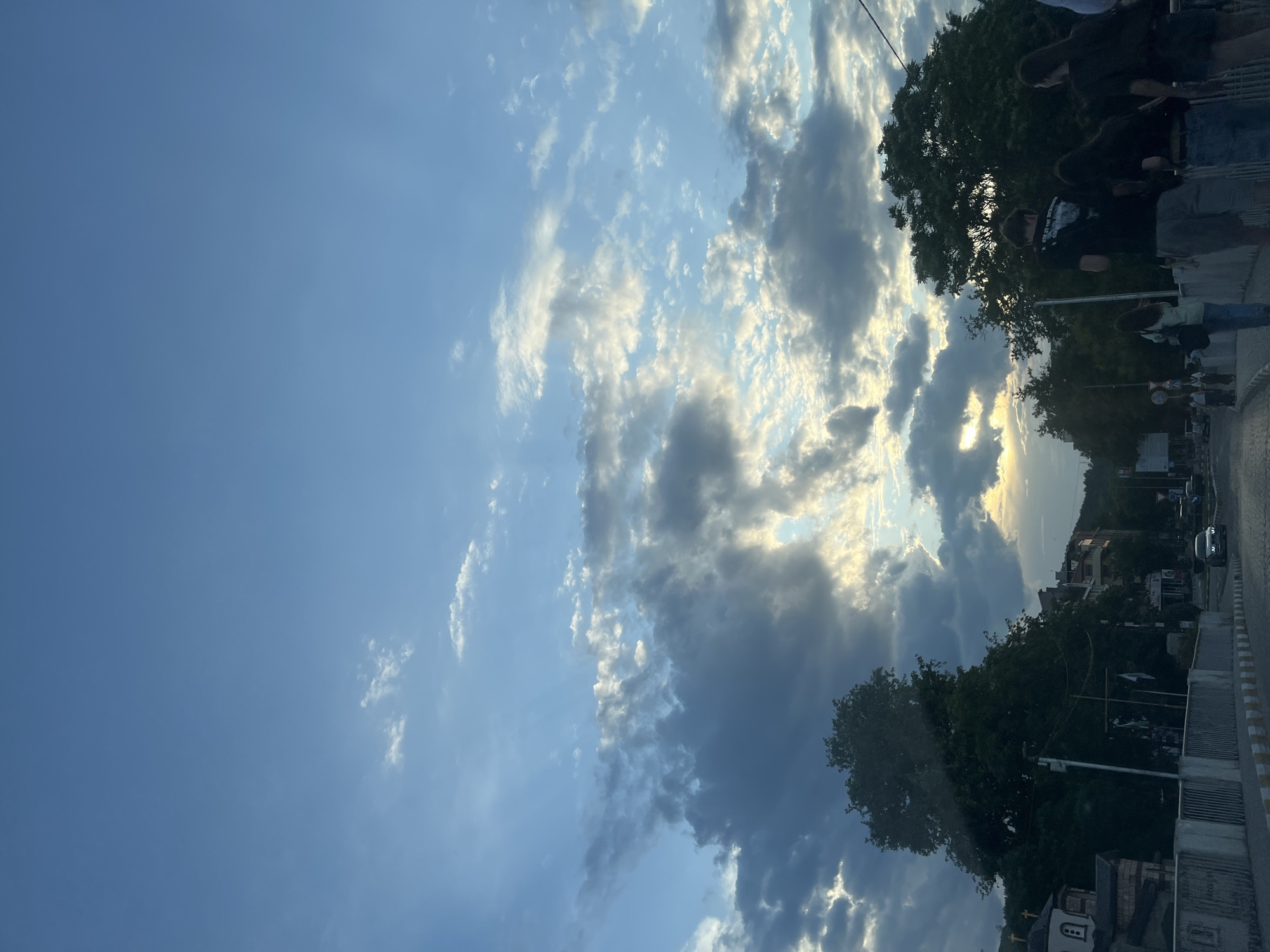 The image depicts an outdoor street scene under a dynamic sky, likely captured during the late afternoon or early evening given the quality of light. The upper and left portions of the frame are dominated by a partly cloudy sky, featuring patches of clear blue, bright white, and darker grey clouds. Distinct sunbeams, or crepuscular rays, are visible breaking through the clouds from the upper right.
In the lower and right sections of the image, a street or road runs horizontally. Lush green trees with dense foliage line the side of the road, partially obscuring buildings behind them. Several individuals are present near the trees and on what appears to be a sidewalk or curb. They are dressed in casual clothing; one person in the foreground wears a dark t-shirt with an illegible white graphic or text design. Further along the street, more buildings of varying architectural styles and at least one vehicle are visible. A circular street sign, typical for traffic regulation, is also discernible amidst the urban elements. The scene conveys a tranquil, everyday moment in an urban or suburban environment, consistent with a street in Asenovgrad, Bulgaria.
