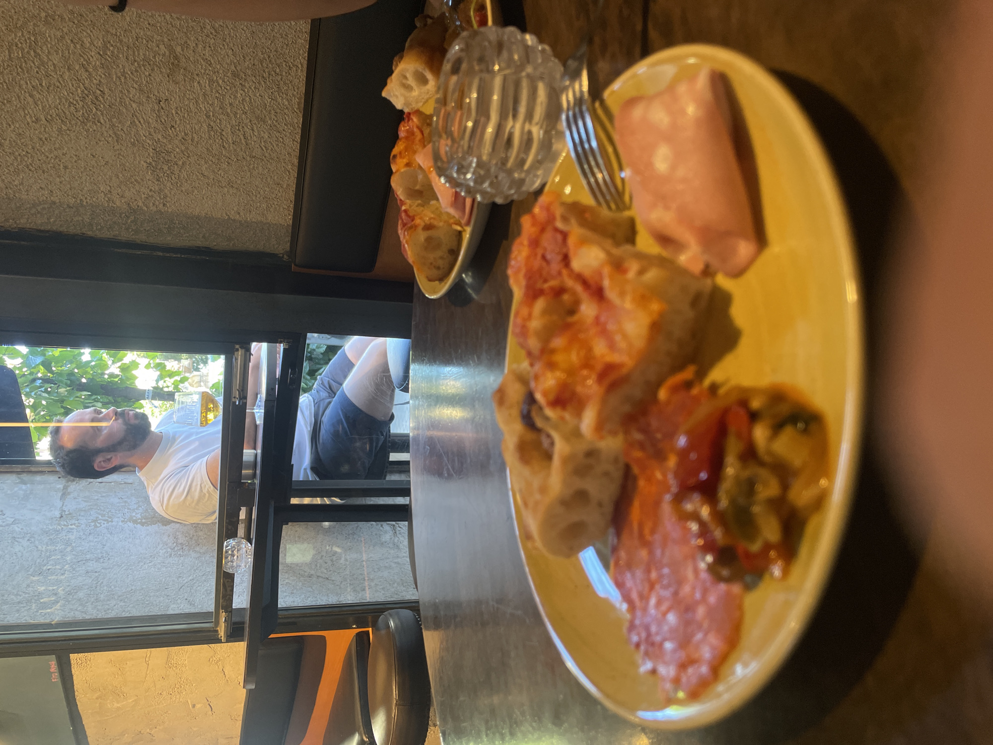 The image depicts a yellow plate of assorted appetizers, including pizza, salami, and ham, placed on a wooden table. Beside the plate is a glass sugar dispenser and a fork. Through a window in the background, a man is visible, wearing a white t-shirt and shorts. He is sitting in what appears to be a chair, enjoying the outdoors. Based on the style of the architecture and general ambiance, the location is likely in Barcelona, Spain.