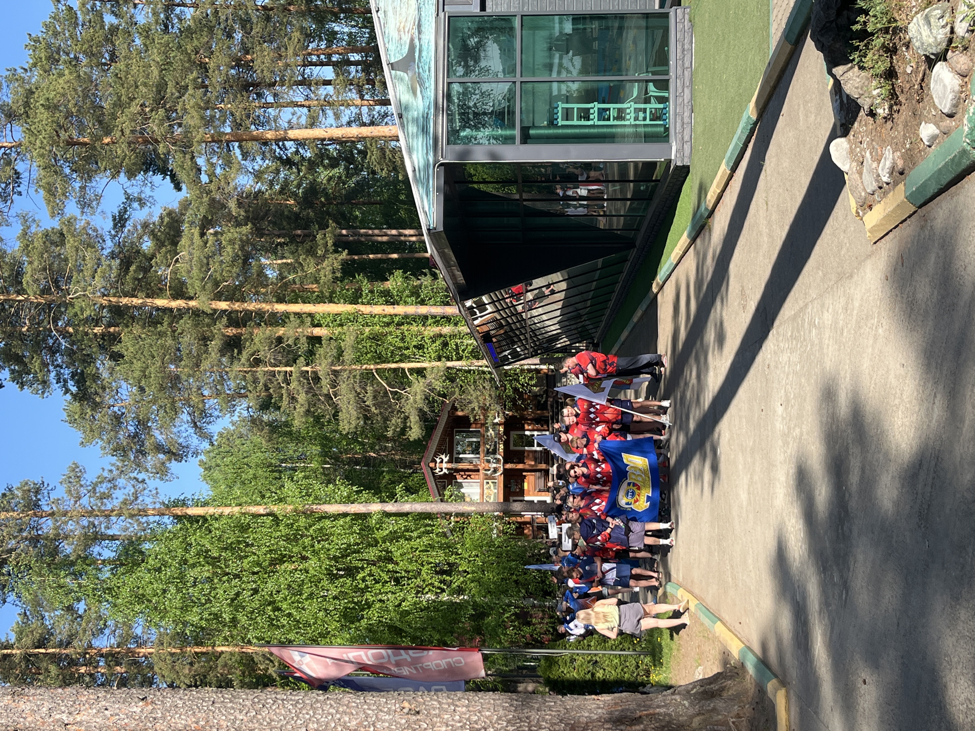 The image shows a group of people standing along a concrete path next to a building and a wooded area. The building has a modern design with large glass windows and a green, grassy border along its base. The path is lined with a curb painted green and yellow.
The group of people appears to be a team, possibly athletes, wearing red and blue jerseys, some holding flags. There are trees in the background.
The location is the ski complex, near the city of Chaykovsky, Russia.