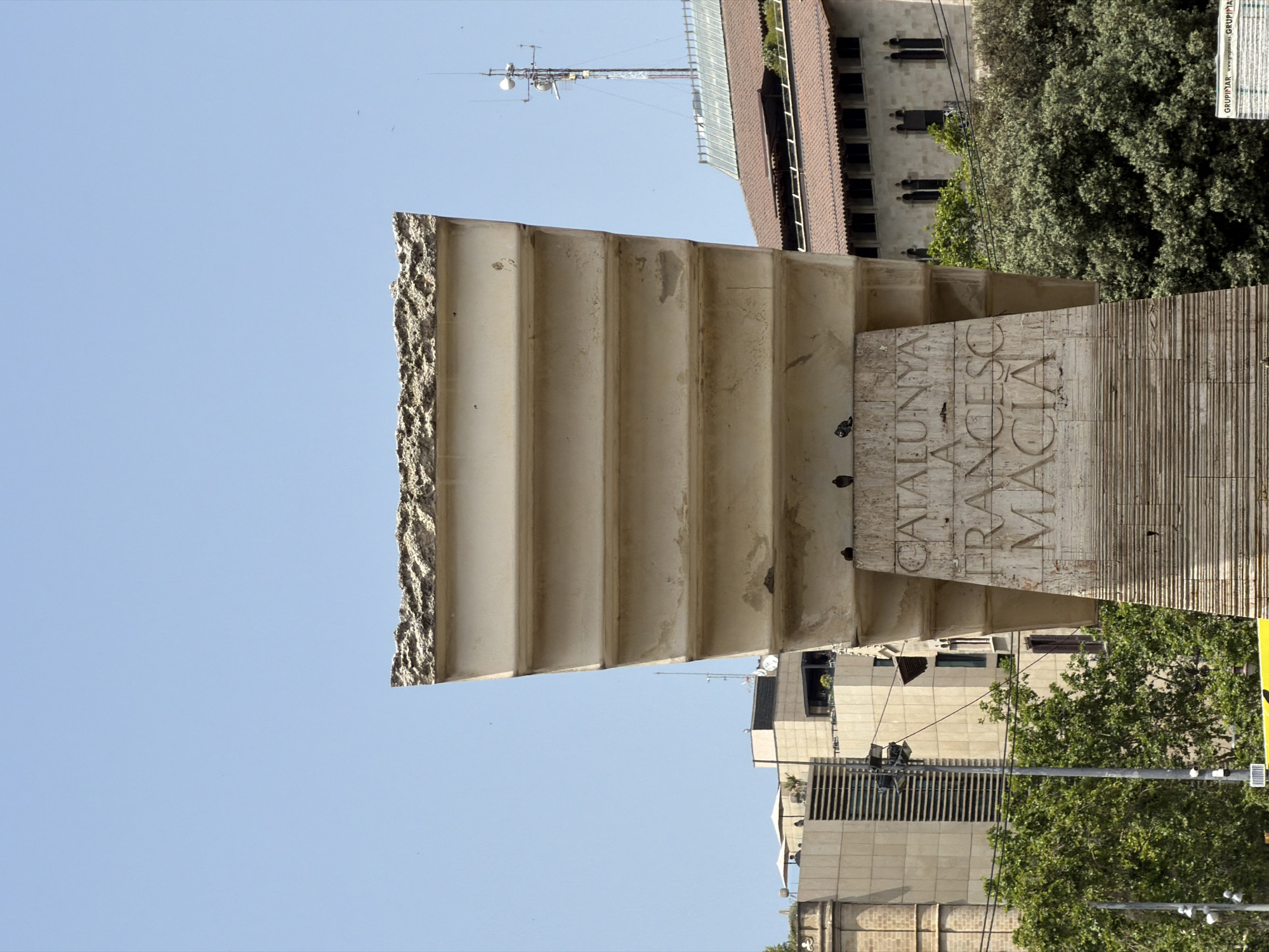 The image depicts a close-up view of a large, weathered stone monument with carved inscriptions, likely in a public space in Barcelona, Spain. The monument is a light beige color and has a tiered, almost architectural design, with horizontal grooved sections and broader, smoother sections where inscriptions are visible. The top of the monument appears to be chipped or broken, revealing a rough texture.
The inscription on the monument reads "CATALUNYA A FRANCESC MACIA," indicating it is dedicated to Francesc Macià, a prominent Catalan politician. The letters are deeply carved into the stone. Several pigeons are perched on the monument, some on the carved inscription and others on the upper tiers.
In the background, several buildings are visible, suggesting an urban setting. To the left, there are modern buildings with large windows and what appears to be a rooftop terrace with umbrellas and people. Further to the right, there's a building with a tiled roof and arched windows, and behind it, a tall communication tower with antennas. Lush green trees are interspersed between the monument and the background buildings, suggesting a park or tree-lined street. The sky is clear and blue. In the bottom right corner, a portion of a sign with text, including the word "GRUPINYAR," is visible. The overall scene suggests a sunny day in Barcelona.
