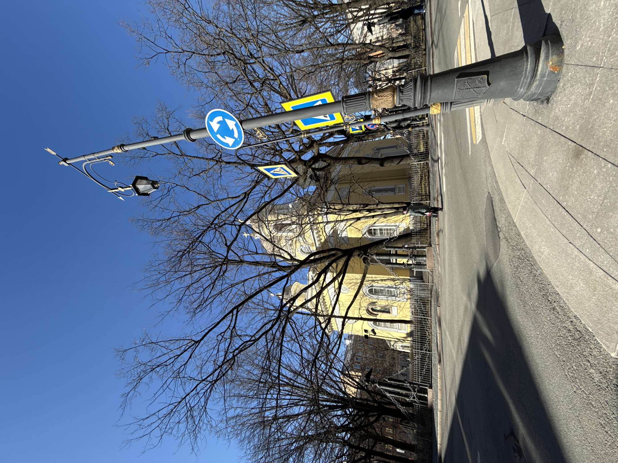 The image shows a street scene in Saint Petersburg, Russia, taken on a sunny day with a clear blue sky. A lamppost with a roundabout traffic sign is in the foreground on the left, along with other traffic signs. Behind the lamppost, a yellow building is visible through bare tree branches, with architectural details suggesting it could be a church or significant historical building. Pedestrian crosswalk stripes are visible on the right, along with a sidewalk. The photograph is taken from a slightly low angle and rotated.