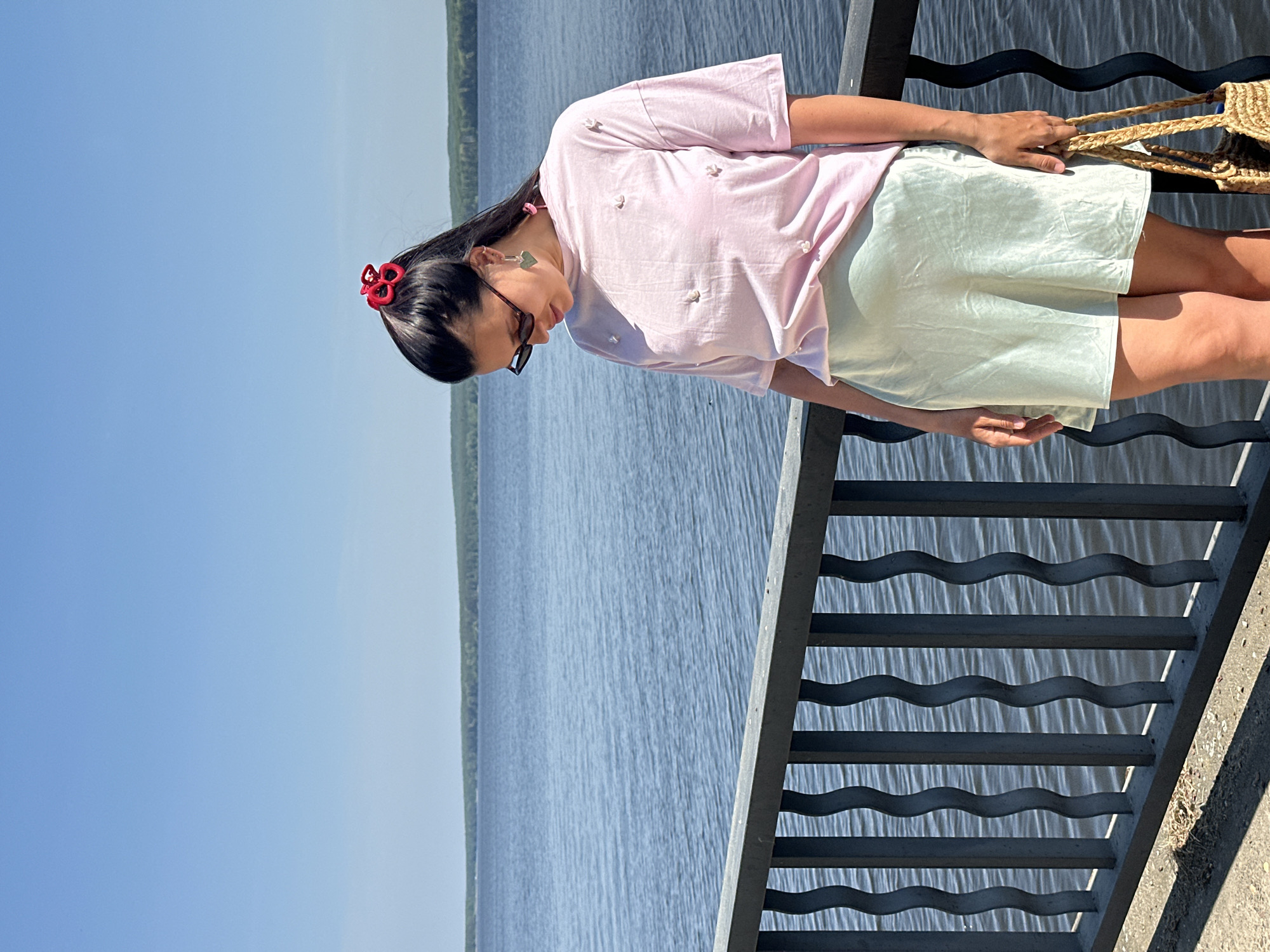 A woman stands on a metal railing platform with a straw bag in Chaikovsky, Russia, on the banks of the Kama River. She wears a pink shirt with floral embellishments and a light green skirt. The backdrop includes a view of the water and trees in the distance.