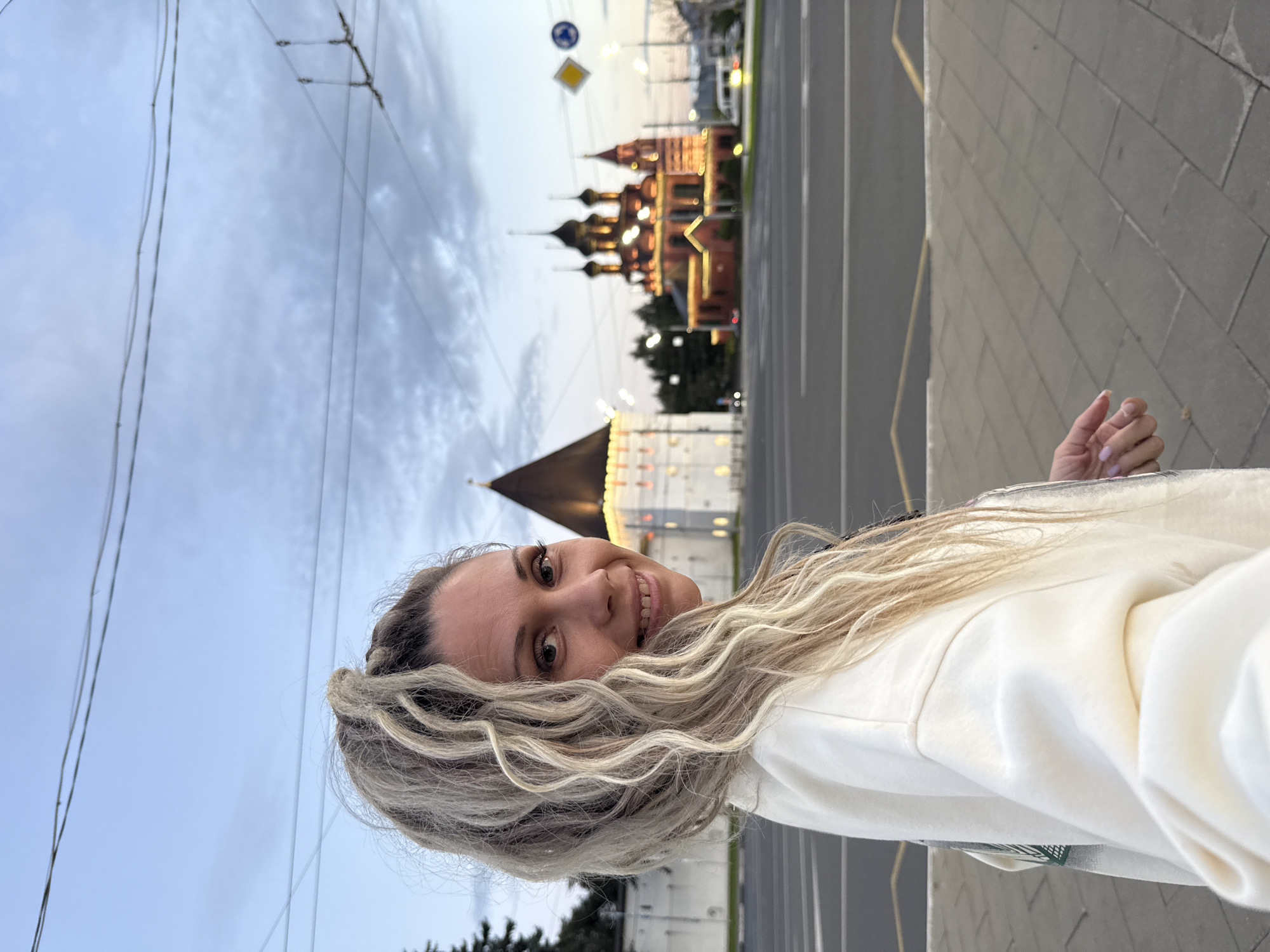 A medium shot captures a Caucasian woman with blonde, dreadlocked hair styled in a high ponytail, looking towards the camera with a slight smile. She is wearing a cream-colored sweatshirt. The background features a street scene in Zhukovo, Russia, with a distinctive red, domed church illuminated by lights and a white tower with a conical roof. The sky is a soft blue with scattered clouds, indicating either early morning or late evening. Overhead, power lines are visible, and a street crossing with yellow markings is in the foreground. The overall atmosphere is calm and urban.