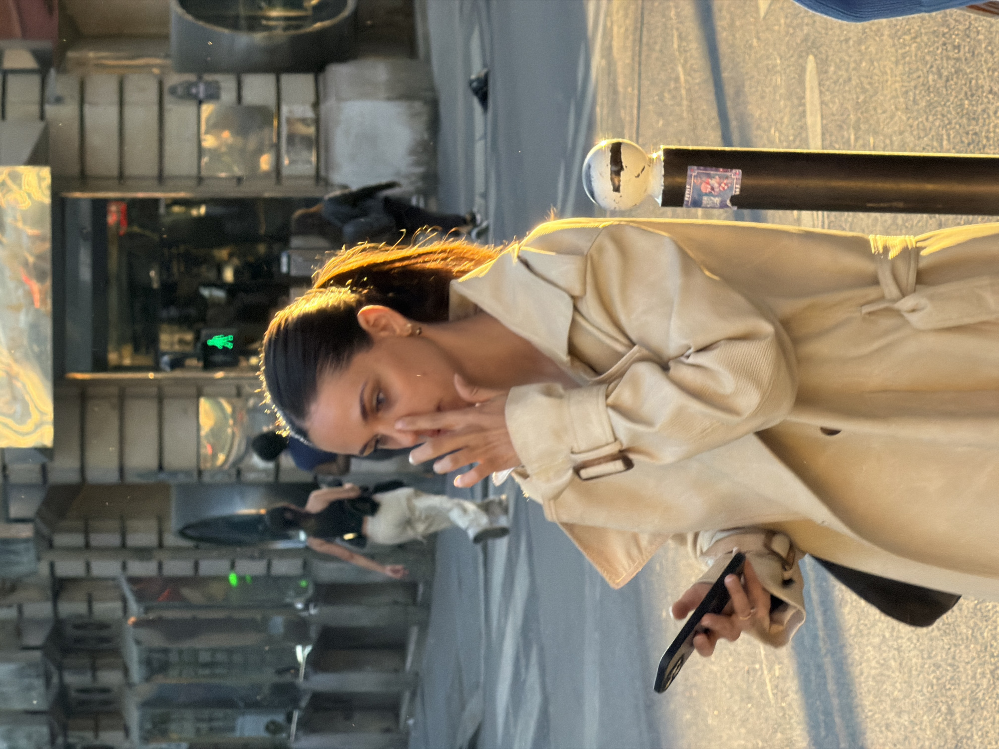 This street photograph, captured in Paris, France, depicts a woman in a candid moment during what appears to be the golden hour, with warm sunlight illuminating the scene. The woman, with dark hair pulled back, is the primary subject. She is wearing a light beige or cream-colored trench coat with the collar and lapels visible, and she has small gold hoop earrings.
She holds a dark-colored smartphone in her left hand, while her right hand is partially covering her mouth and nose, obscuring her exact expression, but suggesting a moment of introspection or a private reaction. Her gaze is directed slightly downwards and to her right.
To the woman's right, a black street pole with a white, spherical top finial is visible. A small, colorful sticker is affixed to the pole, showing a graphic with some blurry text, possibly containing numbers like "1997" or "1994."
The background consists of complex reflections from a large window or glass facade, likely belonging to a building or shop. These reflections show blurred architectural elements and other indistinct figures. Notably, within the reflections, a green neon sign in the shape of a crocodile or alligator is visible, strongly suggesting the Lacoste brand. Another faint reflection shows a person in light pants and a dark top further in the background. The overall setting is an urban street, characterized by bright light and reflections.