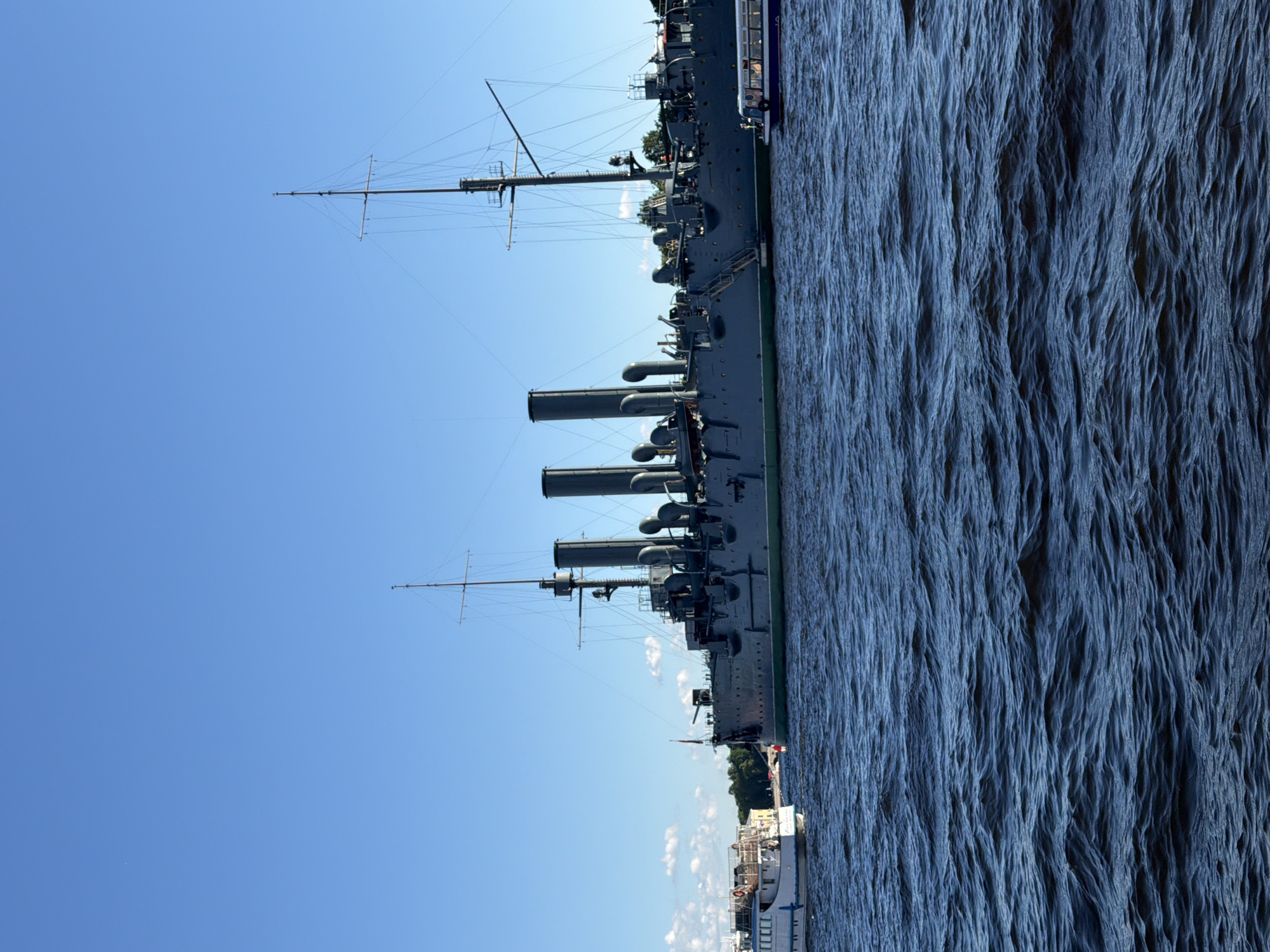The image depicts a wide shot of the Aurora cruiser, a historical Russian protected cruiser, docked on the Neva River in Saint Petersburg, Russia. The ship, a museum ship and a symbol of the Russian Revolution, is prominently featured in the mid-ground. It is a large, grey vessel with multiple smokestacks and masts. The water in the foreground is dark blue and choppy, indicating a breezy day. To the left of the Aurora, a smaller white passenger boat with a red stripe is visible, also on the water. In the far background, the distant bank of the Neva River can be seen with trees and buildings, hinting at the cityscape of Saint Petersburg. The sky above is a clear, bright blue with a few scattered white clouds, suggesting fair weather. The scene is captured from the perspective of someone on another boat or the riverbank.
