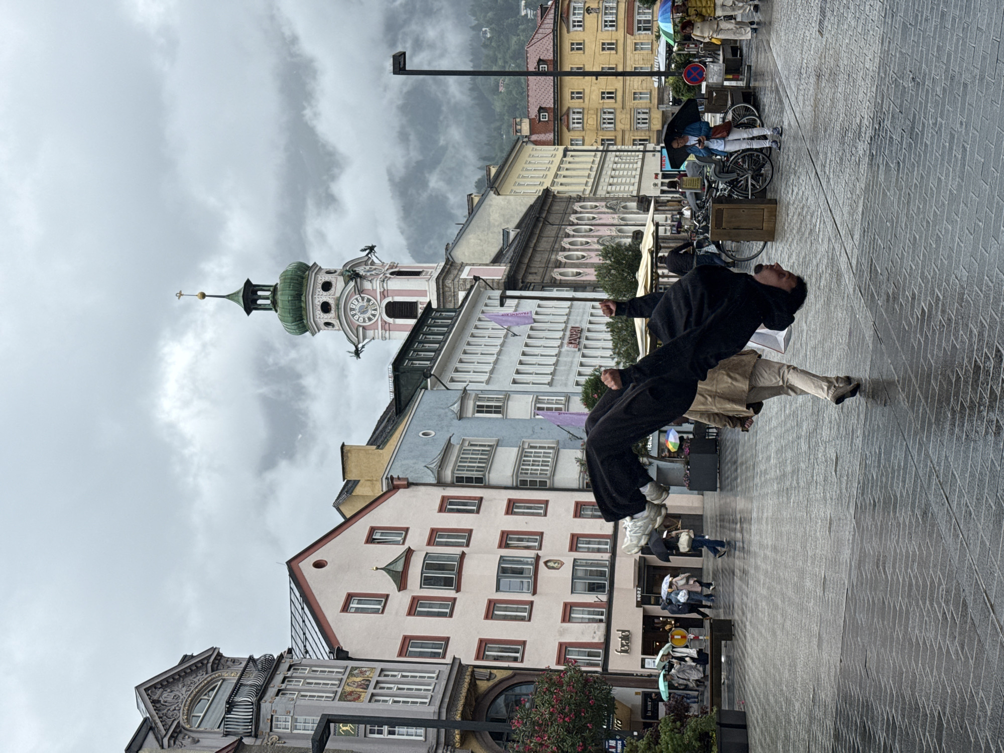 An athletic individual performs a flip in mid-air in the middle of a public square. The person, dressed in black pants and a black hoodie, is upside down with their legs extended. They are in the foreground, with a blurred background of people walking with umbrellas and colorful buildings. In the background, a tall clock tower with a green dome stands out. The ground is wet and reflects the surroundings, indicating it is raining or has recently rained. The architecture suggests the location is likely Innsbruck, Austria, given the distinctive clock tower of the city's Old Town Hall, also known as the "Stadtturm". The sky is overcast and cloudy.