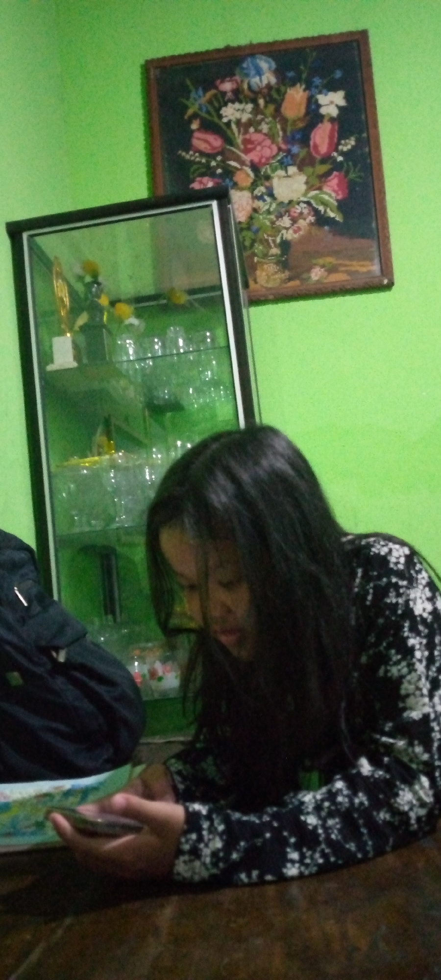 The image depicts an indoor scene with a young female in the foreground, seated at a wooden table and looking down at a mobile device she is holding in her hands. She has long, dark hair and is wearing a black and white patterned long-sleeved shirt. To her left is a black backpack or bag. In the background, a glass-fronted display cabinet filled with clear glasses and some decorative items, including a trophy, is visible. Above the cabinet, a framed tapestry or artwork depicting a bouquet of flowers hangs on a bright green wall. The overall impression is that of a casual, domestic environment, possibly a living room or dining area in a home in Sumberwono, Indonesia. The lighting is dim, casting shadows and contributing to a slightly muted color palette.