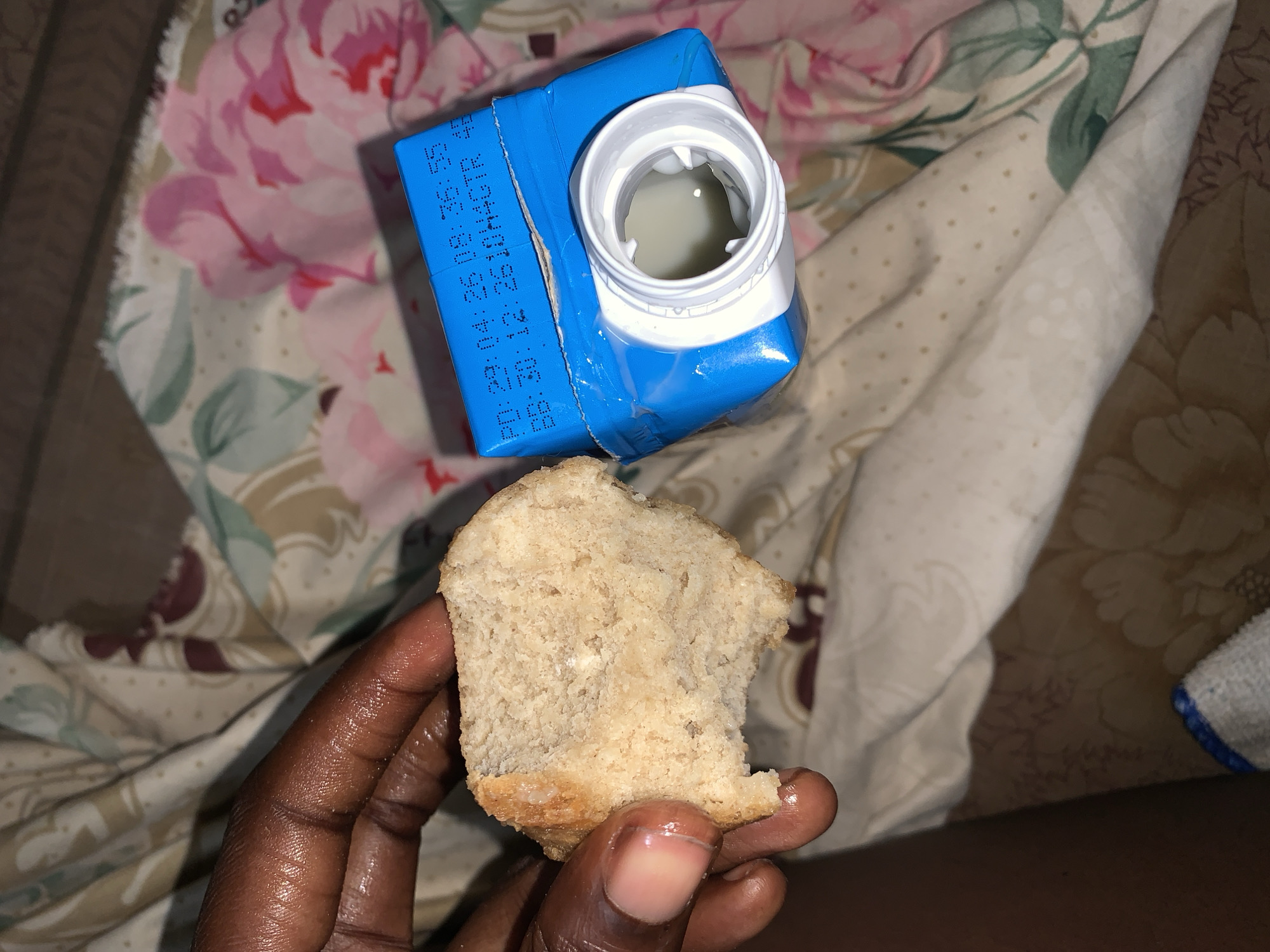 The image provides a close-up view of a person's dark-skinned hand holding a substantial piece of light brown bread, which appears to have a dense, slightly moist crumb and a visible crust at the bottom. The bread looks as if it has been broken off from a larger loaf. Adjacent to the hand, on a patterned fabric surface, rests an open blue carton, likely containing milk or a similar white liquid, which is visible inside the white screw-on cap.
Black text is printed vertically on the side of the blue carton. Clearly visible is "BB: 29.04.18 08:35 S", with additional partially visible lines that seem to include "26. JJAAGTR" and "TR: 0:13". These numbers likely indicate a best-before date and possibly a production batch code or time.
The setting appears to be indoors, with various soft fabrics serving as the background. These include a fabric with pink floral and green leaf patterns, and another with subtle light dots. A small part of a white and blue towel is also visible on the right side of the frame. The overall lighting suggests a dim environment, possibly early morning or evening. The scene, set in Rafin Par, Nigeria, depicts a simple, solitary meal or snack of bread and a beverage. No specific emotions are discernible.