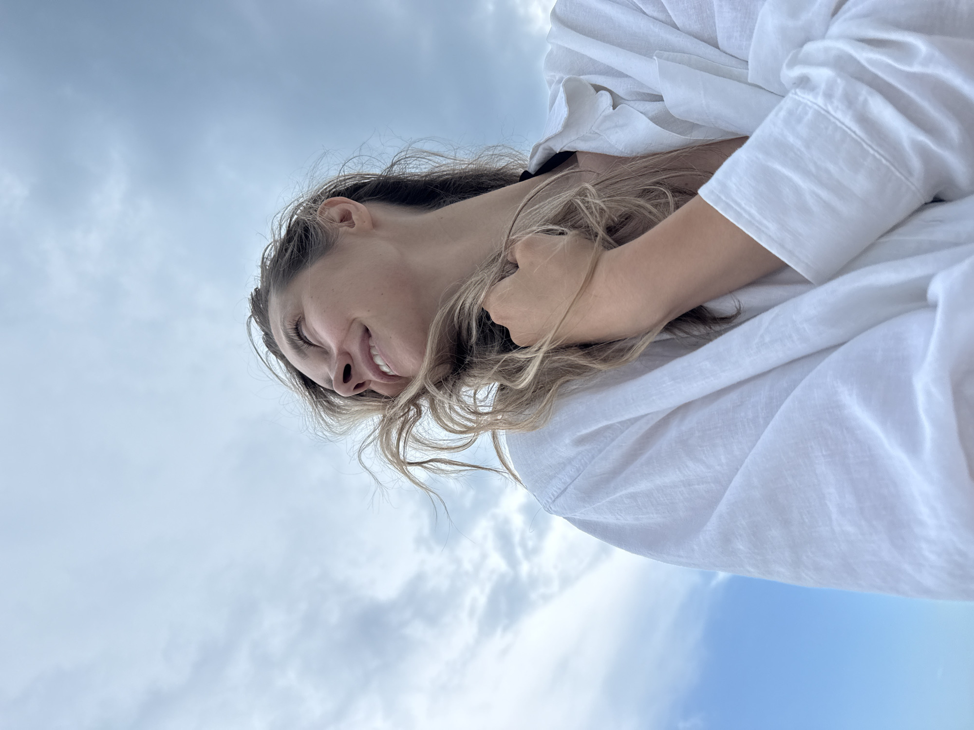 An outdoor, low-angle, medium shot shows a young woman with blonde hair and fair skin, wearing a white collared shirt, looking up and to her right with a joyful expression and a slight smile. Her right hand is tucked into her hair, and her hair is blowing to her left. The background is a cloudy sky. The image was taken in Nebug, Russia.