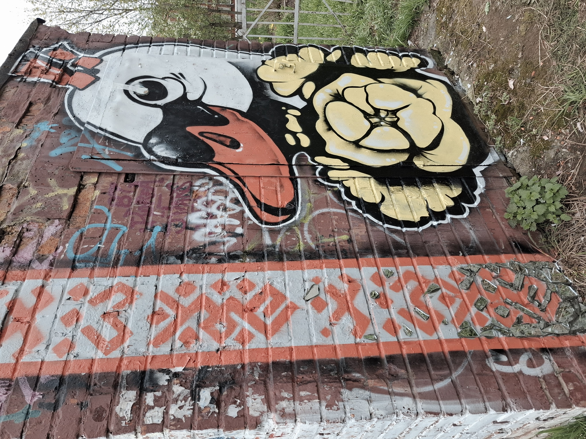 A weathered brick wall, presumably in Glazov, Russia, is covered in graffiti. On the upper part of the wall, there is a large cartoon-like image of a bird's head with an open beak next to a flower. Below the bird and flower, a horizontal band of gray paint is decorated with a repeating orange geometric pattern. The lower section of the wall shows further deterioration and accumulated discoloration, with additional faded spray paint. There is an area on the right-hand side that has broken glass glued onto the wall. A small tuft of vegetation is visible in the upper right corner where the wall meets the ground. The overall impression is of neglected urban infrastructure marked by vandalism and weathering.