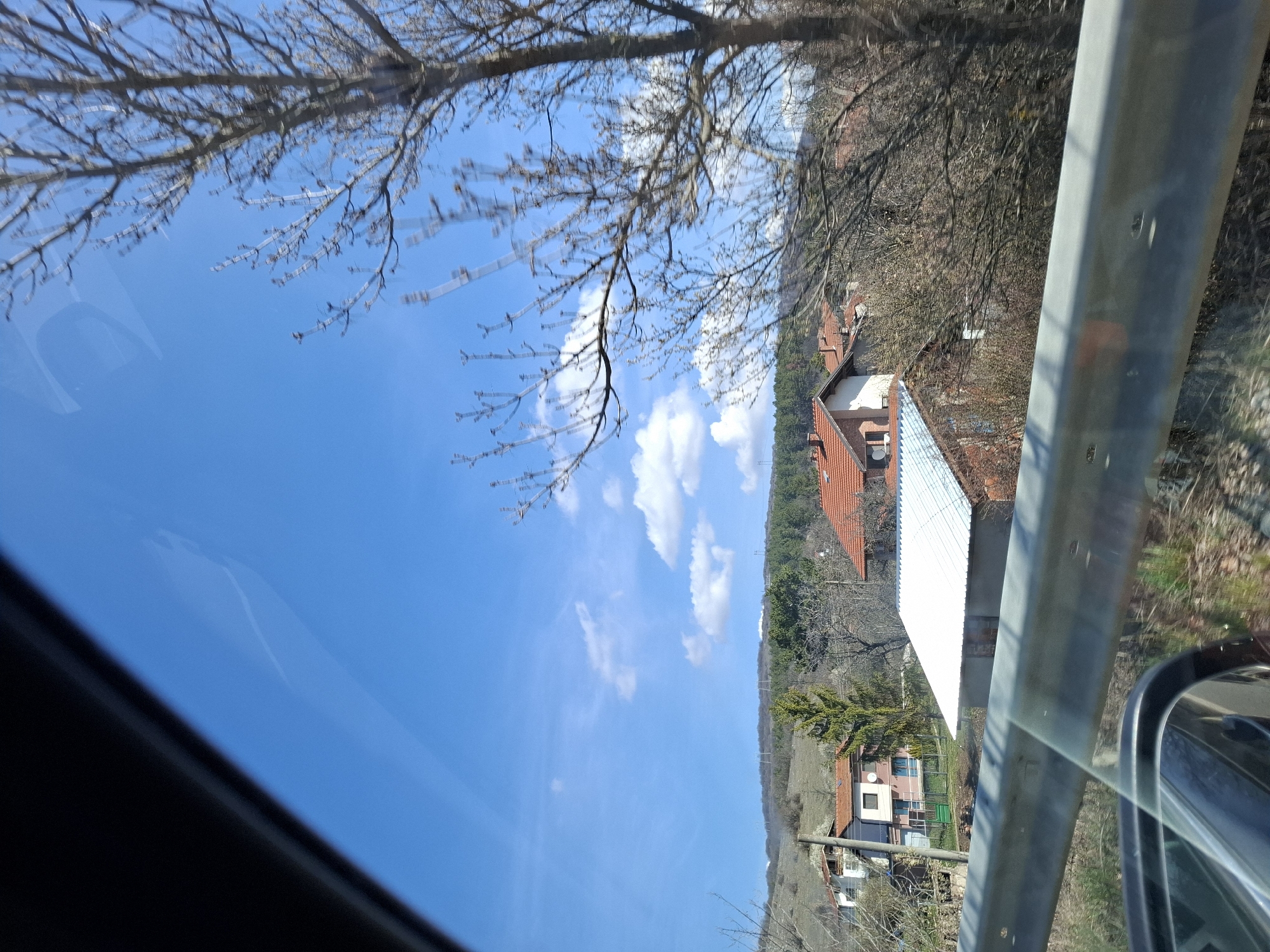 The image depicts a view from inside a vehicle, likely a car, looking out over a landscape near Gradets, Bulgaria. The sky is clear blue with scattered white clouds. Bare tree branches fill the upper left portion of the image, obscuring the sky slightly. In the mid-ground, there are several buildings with orange tiled roofs and white walls. These are set against a backdrop of a forested area and a gently sloping hillside. A guardrail is prominently visible in the right portion of the image, as well as a side view mirror of the car. The vegetation appears to be dormant or in early spring, with many bare trees and brownish undergrowth.