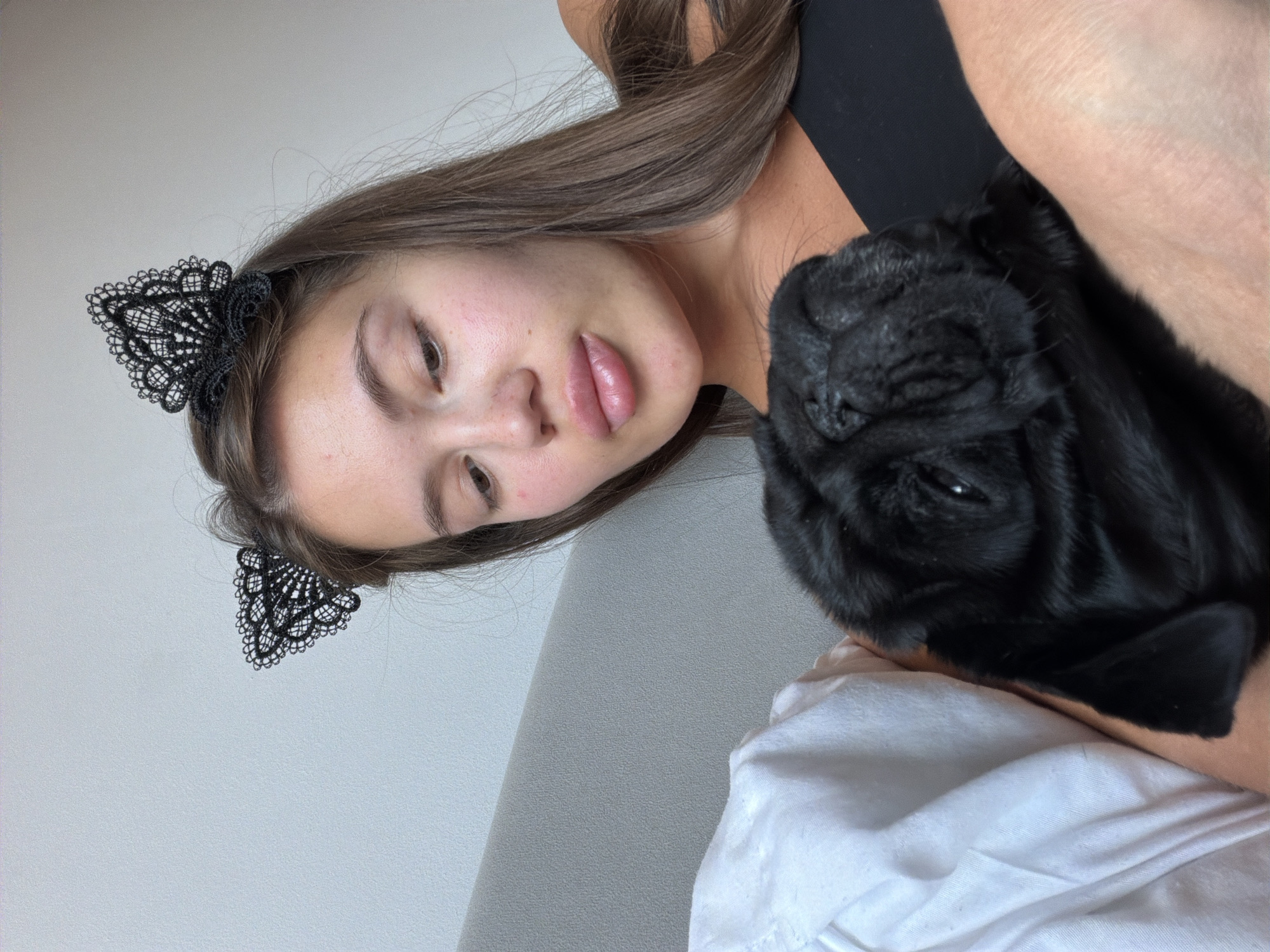 The image shows a close-up of a young woman with long, brown hair and a black lace headband shaped like cat ears. She is wearing a black top. Next to her, resting on her shoulder and chest, is a black pug. The pug's face is partially obscured, but its dark fur and wrinkled features are visible. The woman's face is turned towards the pug, and her expression appears soft and affectionate. The background is a plain, light-colored surface, possibly a wall or furniture, with a hint of a white fabric, perhaps a blanket or clothing, in the foreground. The lighting suggests it is daytime, with natural light illuminating the subjects. The overall mood is intimate and tender. There is no visible text in the image.
