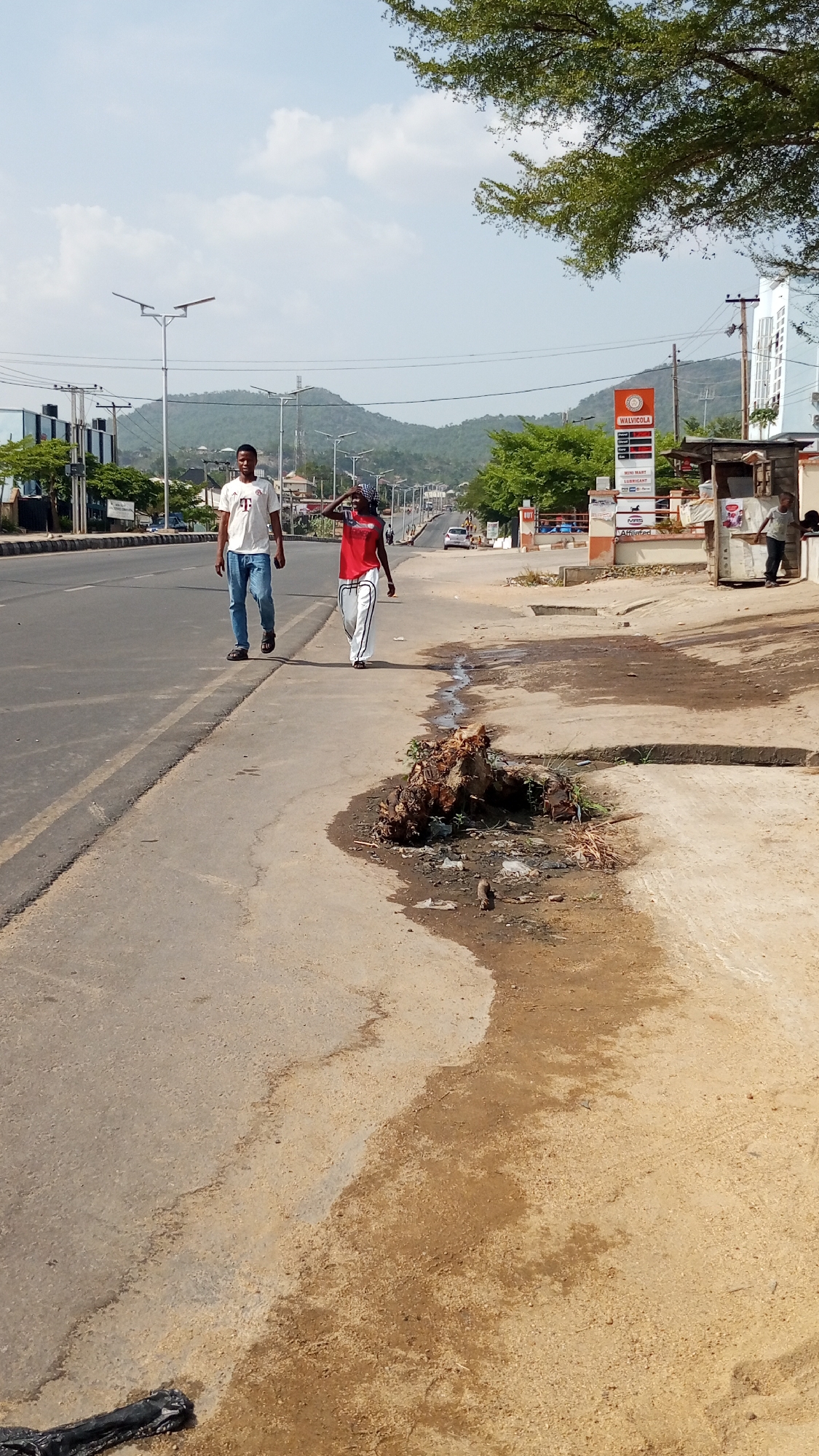 The scene is a street in Adankolo, Nigeria, showing two people walking alongside a road with a Walvicola gas station in the background. One person is wearing a white t-shirt and blue jeans, and the other is wearing a red shirt and white pants, with a headscarf. The road is wide with a dividing line. On the right side of the road, there is a large pile of dirt and debris. A man stands near a small structure to the right of the gas station. The backdrop features hills and utility poles. The weather appears sunny.